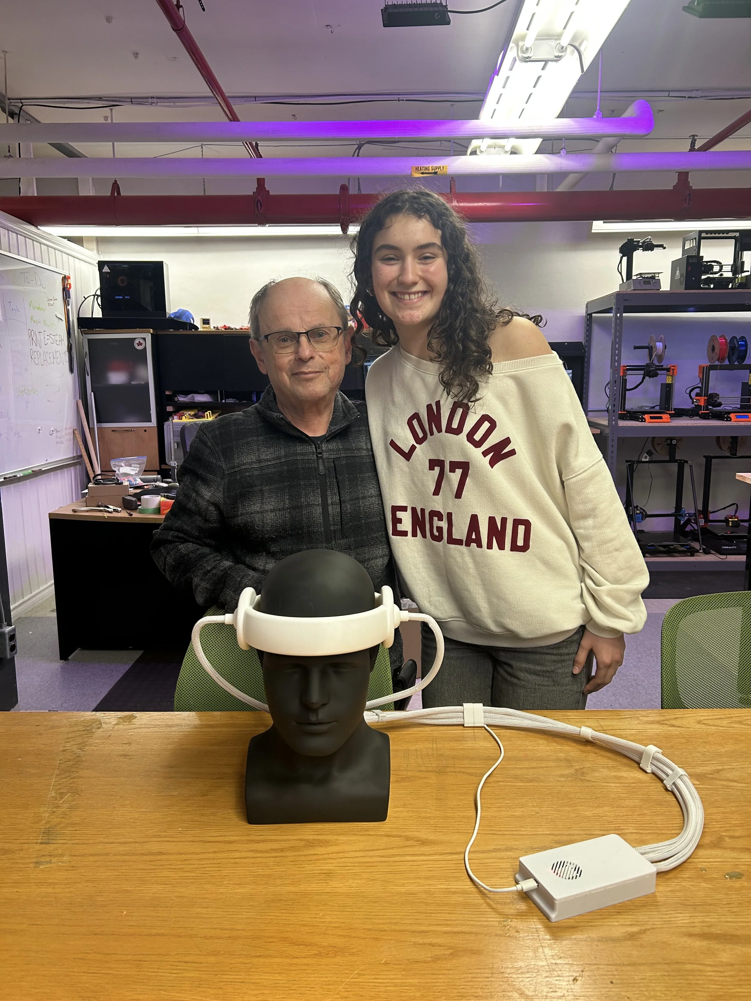 A man and a young woman standing together in a workshop, smiling for the camera. The man is wearing glasses and a dark jacket, while the young woman is dressed in a light-colored sweatshirt with 'LONDON 77 ENGLAND' written on it. In front of them on the table is a mannequin head with a device resembling a brainwave or EEG cap connected to a small white box, with cables running from the cap to the box.