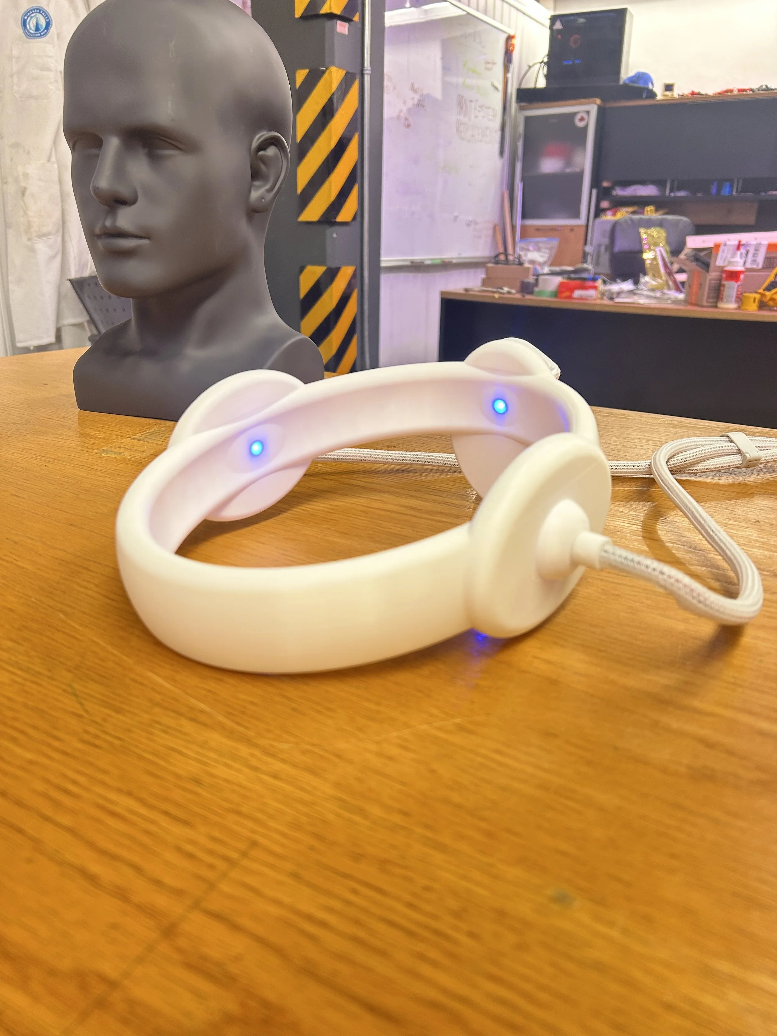 A white headset with blue indicator lights on a wooden surface, with a black mannequin head bust and a cluttered workshop in the background.