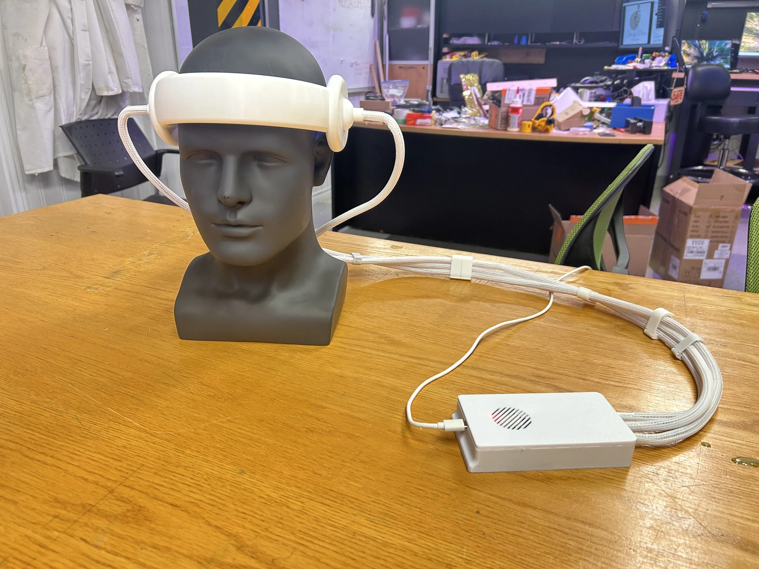 A mannequin head on a wooden table with a white headset connected to a white electronic box. The background shows an office or workshop with various tools, supplies, and computers.