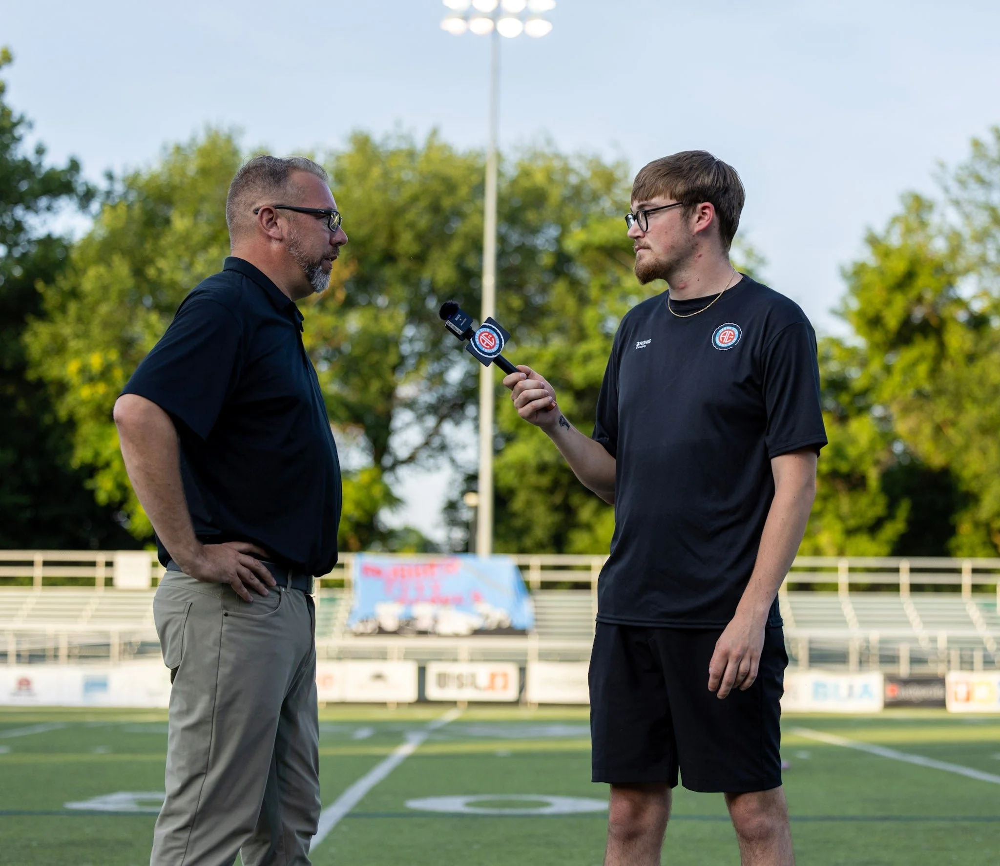 A man in a black shirt being interviewed by a young man holding a microphone on a football field with trees and a blue sky in the background.
