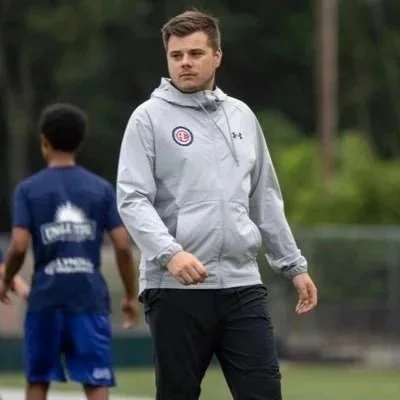 A man in a gray hoodie walking on a sports field with a child in sports attire in the background.