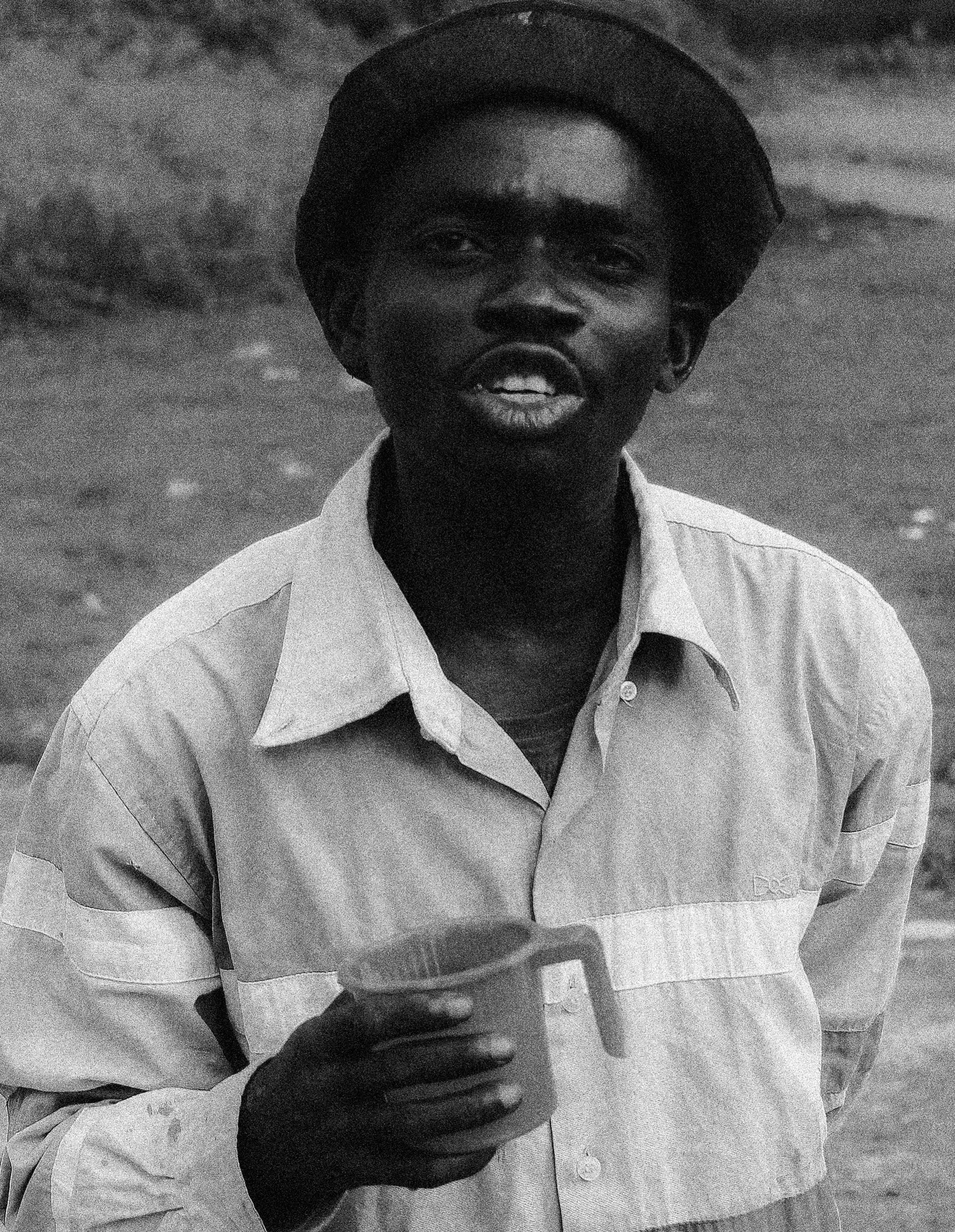 A man with dark skin, wearing a wide-brimmed hat and a light-colored shirt, holding a mug, outdoors, Photographed by Ussi'n Yala in Lambarene, Gabon, 2016