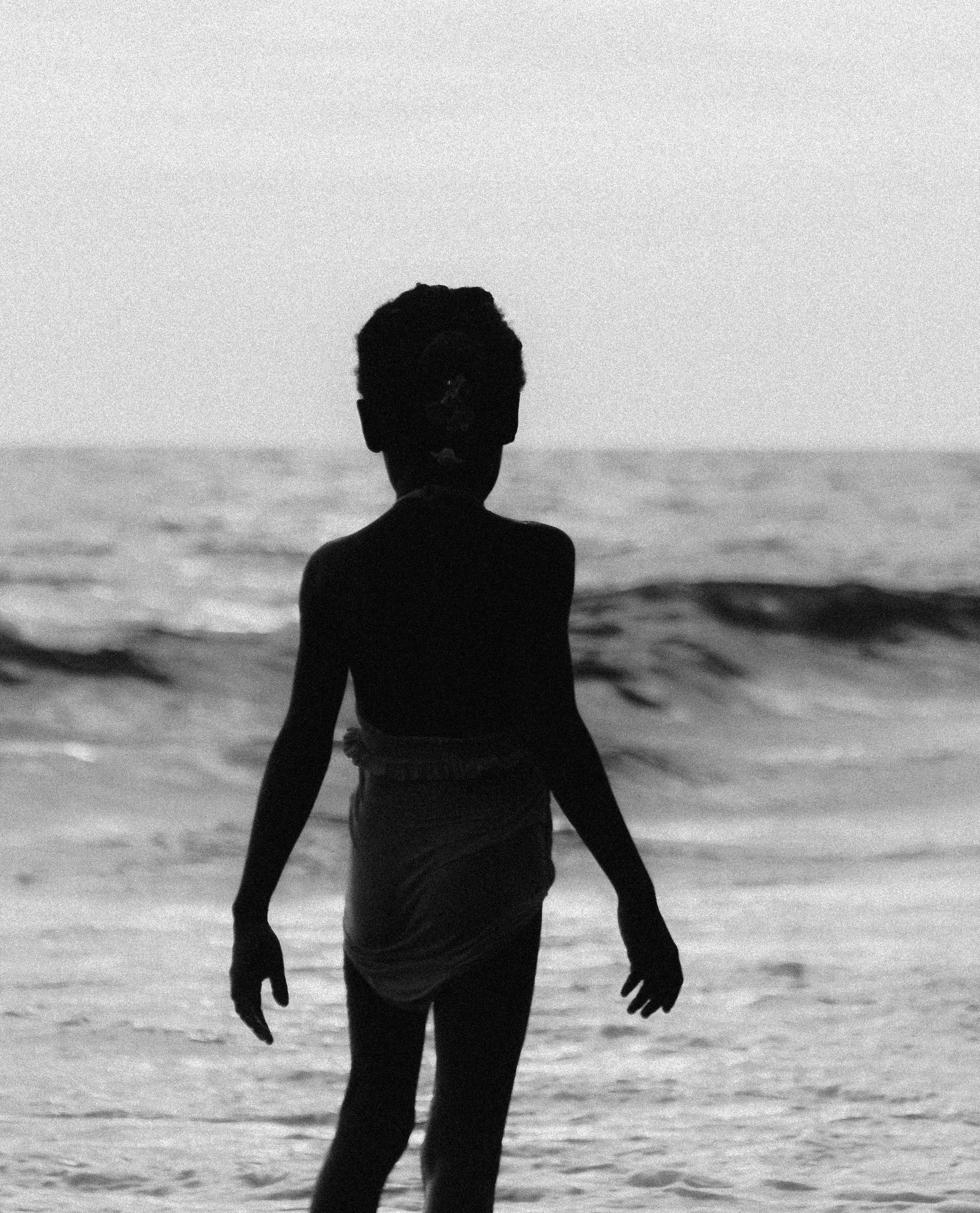A silhouette of a young person standing on the beach near the ocean with waves in the background, Photographed by Ussi'n Yala in Libreville, Gabon, 2016