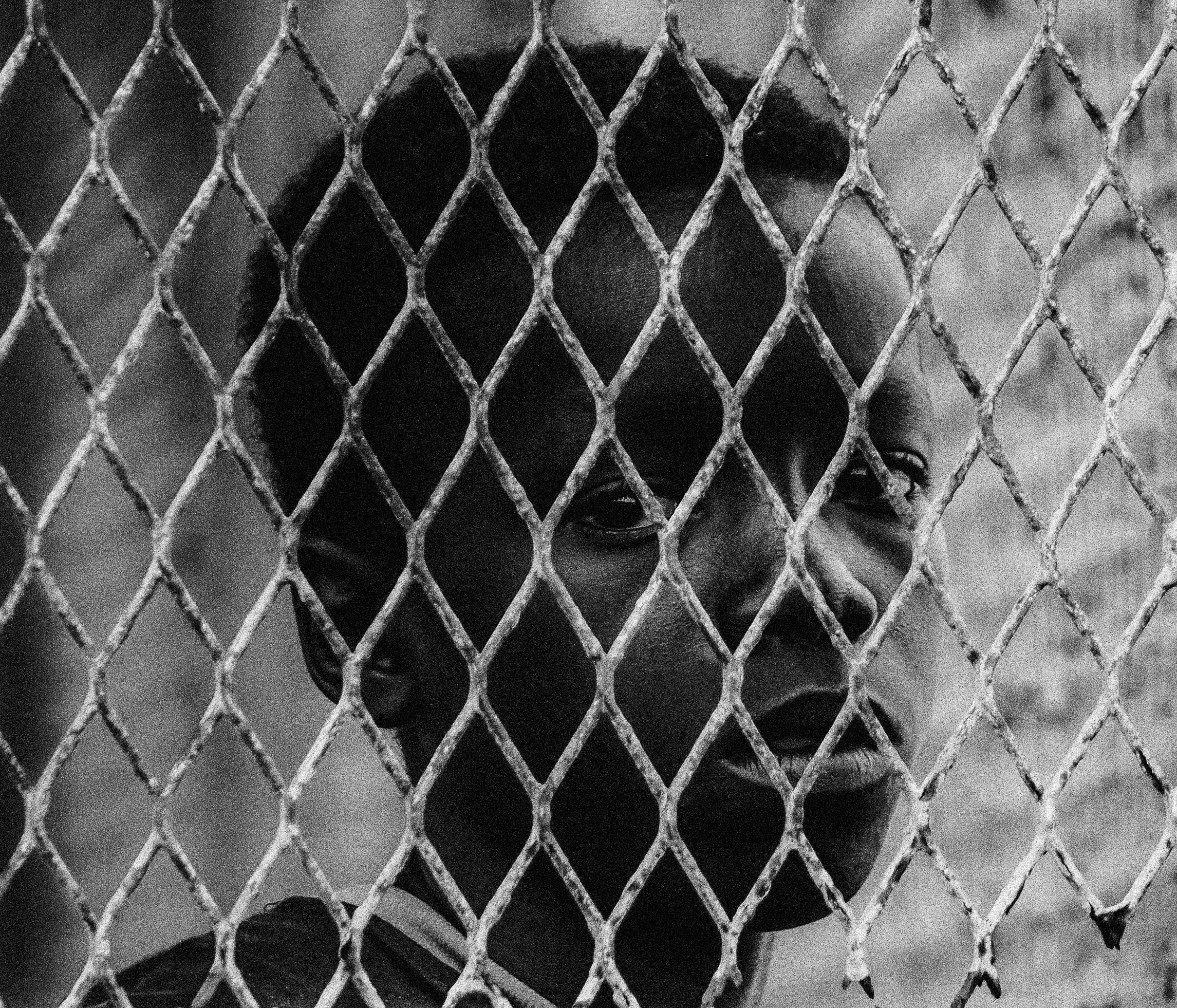 A black and white photograph of a young boy looking through a chain-link fence with a serious expression, Photographed by Ussi'n Yala in Gamba, Gabon, 2016