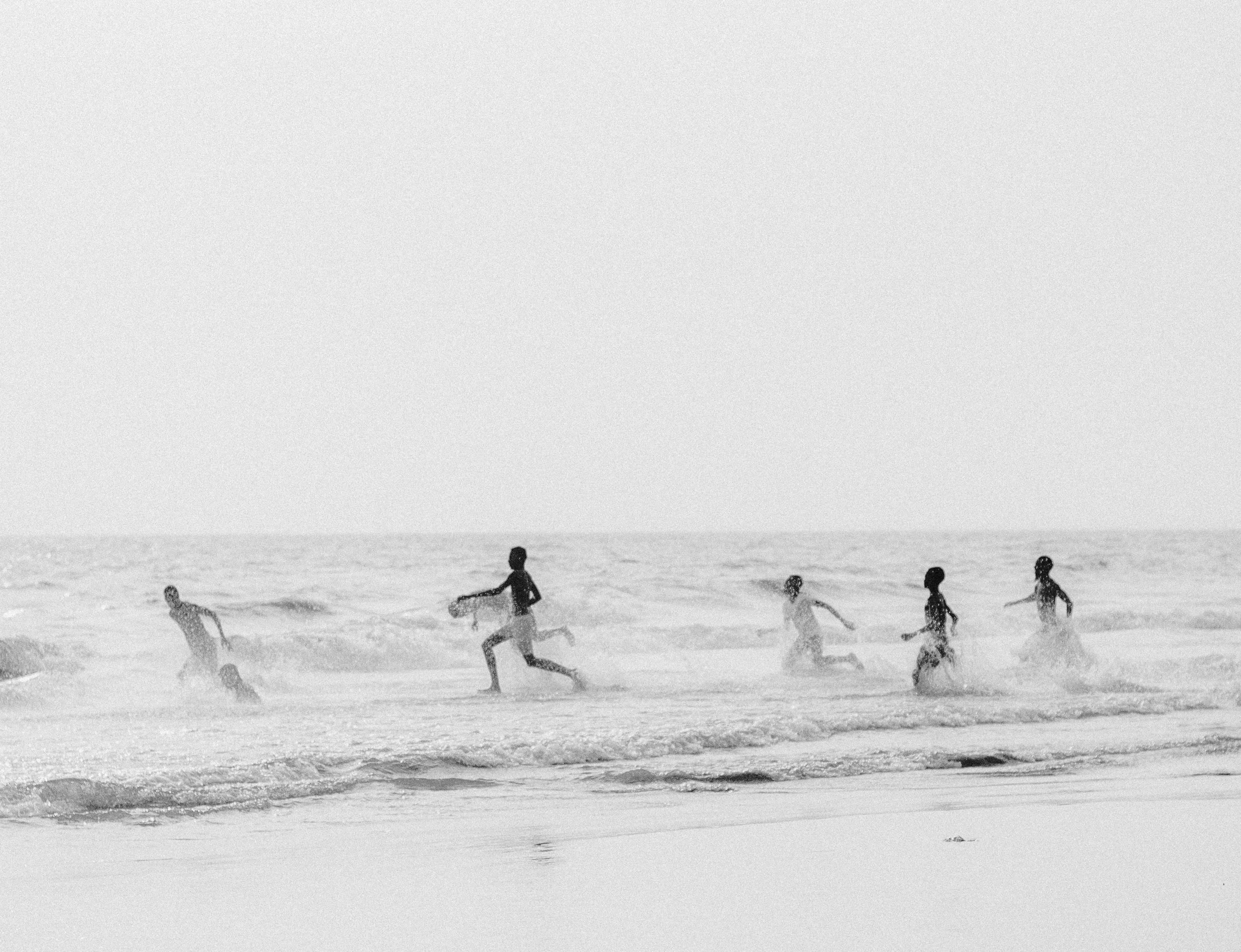 Black and white photo of six children playing and running in the ocean waves at the beach, Photographed by Ussi'n Yala in Port-Gentil, Gabon, 2016