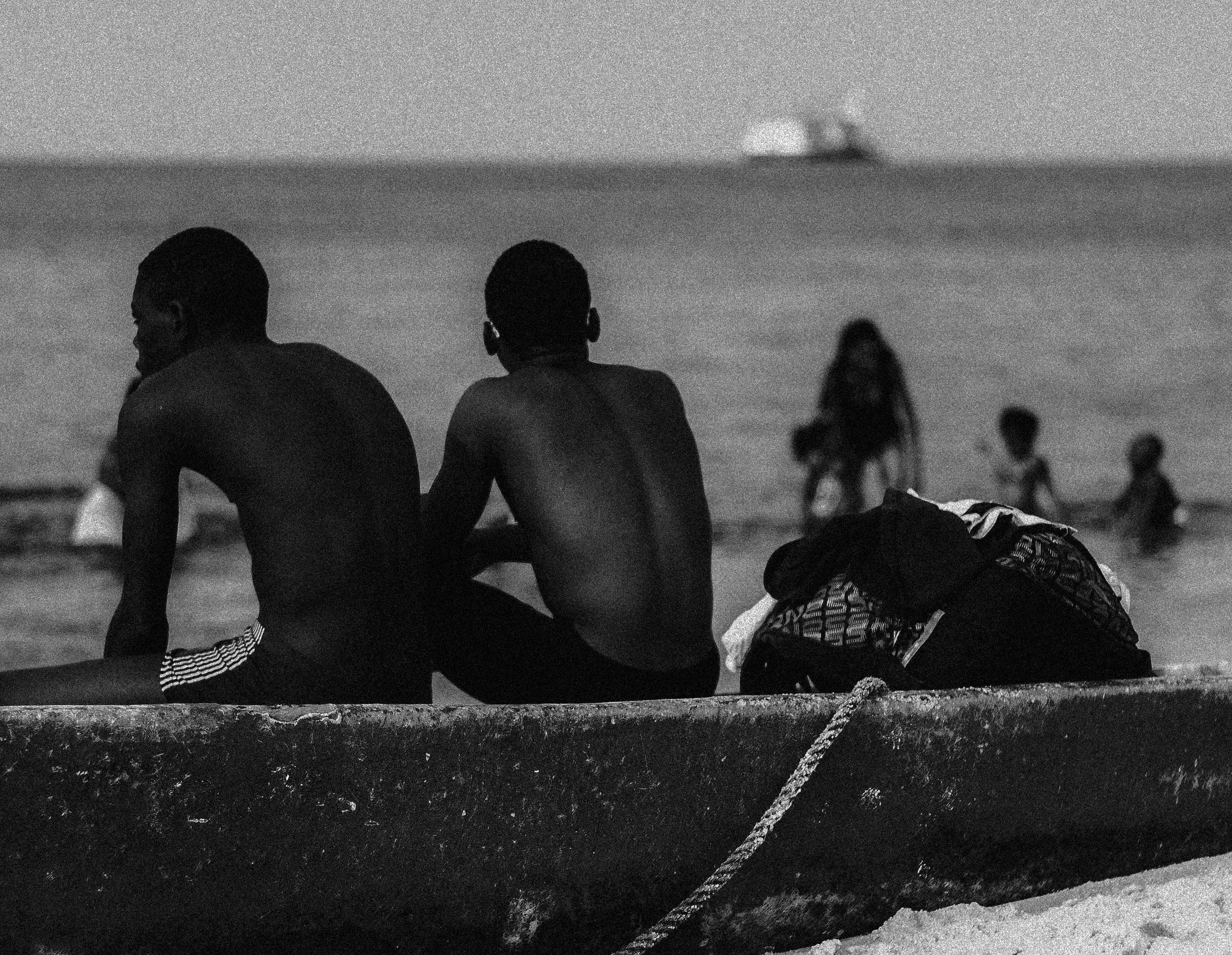Two shirtless boys sitting on a boat by the beach, with a backpack and a rope in the foreground, and a woman and children in the water in the background, Photographed by Ussi'n Yala in Port-Gentil, Gabon, 2016