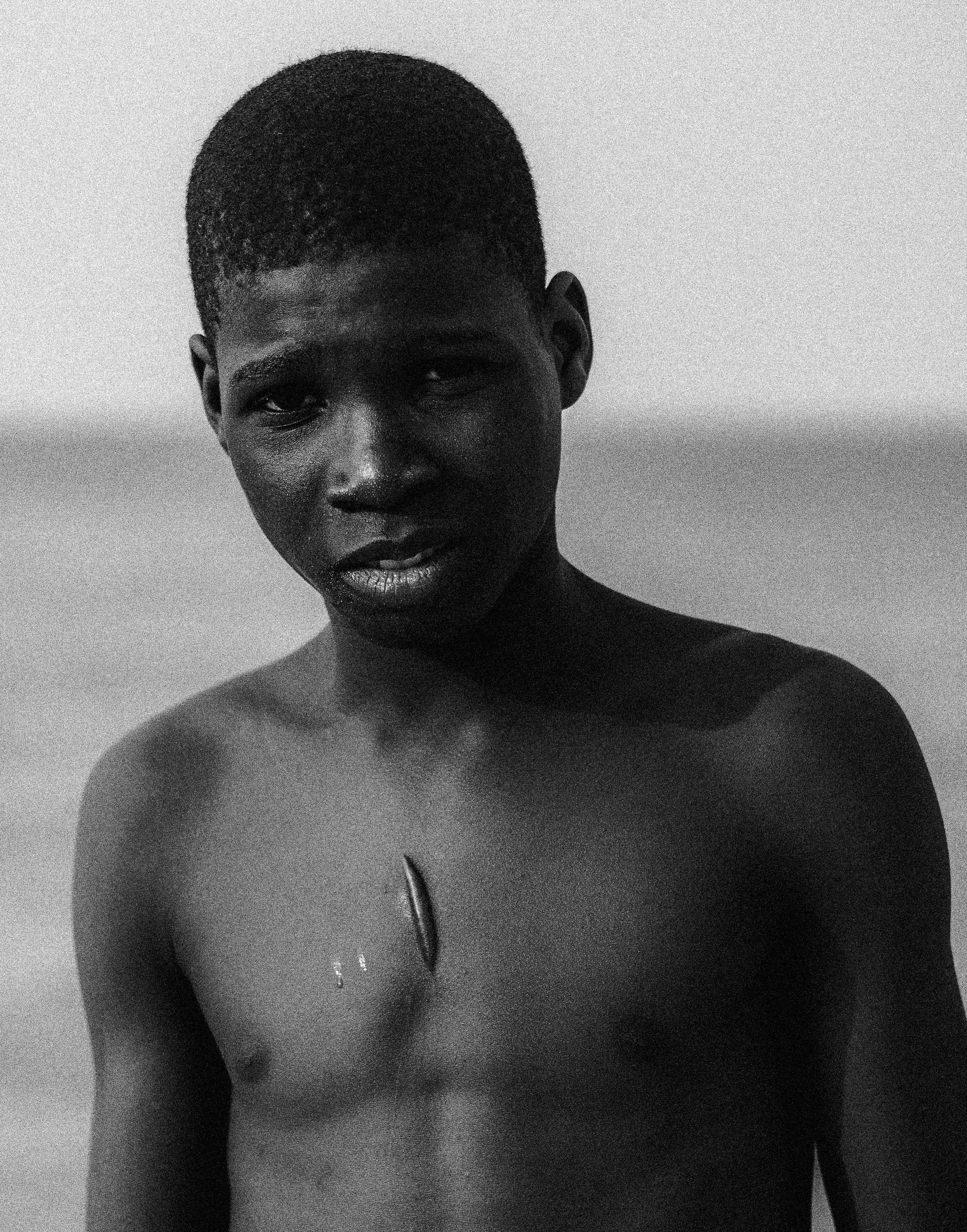 Black and white photo of a shirtless young Black boy with short hair, standing against a plain background, Photographed by Ussi'n Yala in Port-Gentil, Gabon, 2016