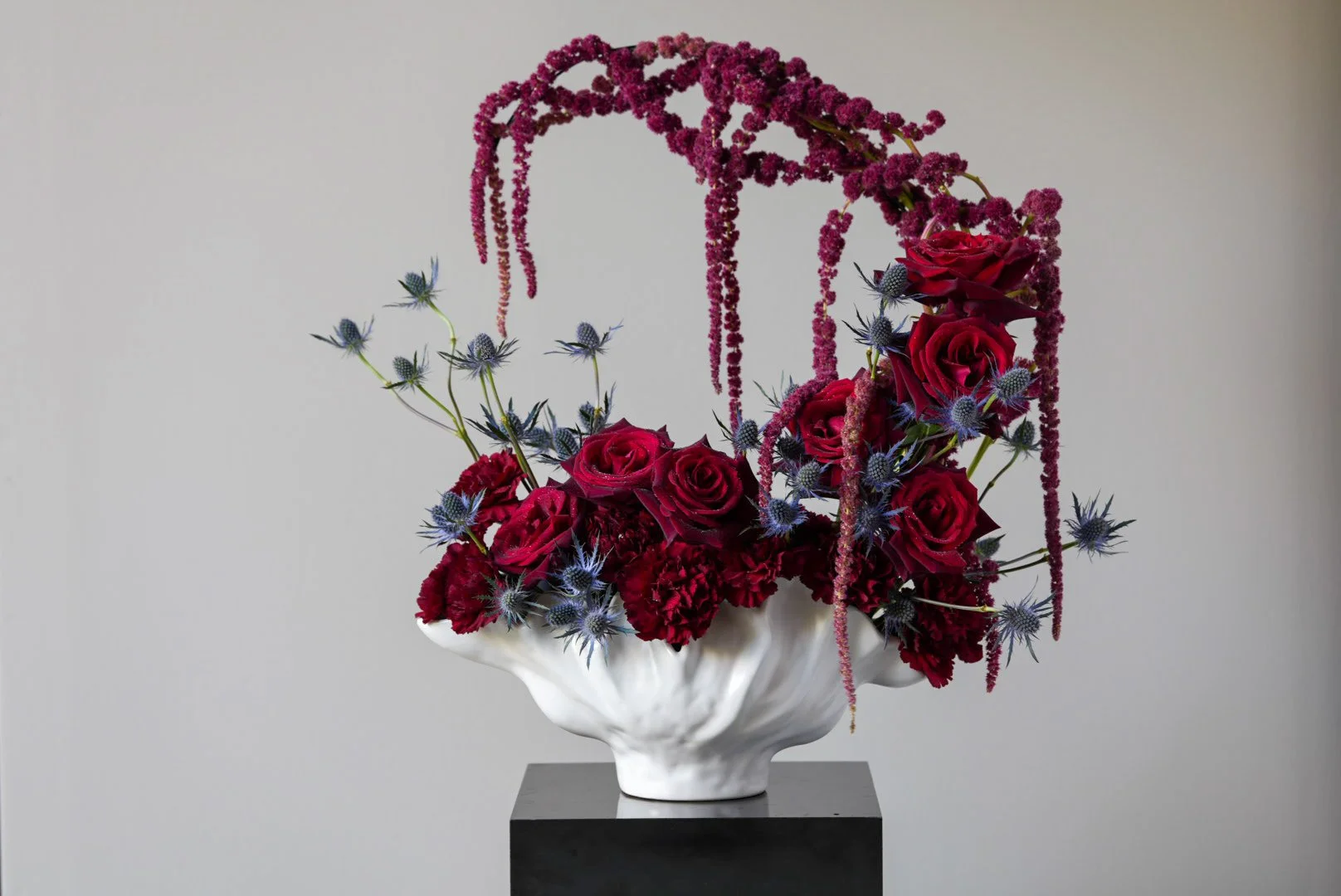 A sculptural flower arrangement in a white, seashell-shaped vase contains deep red roses, red carnations, purple thistles, and trailing pink amaranth, displayed on a black pedestal against a plain background.