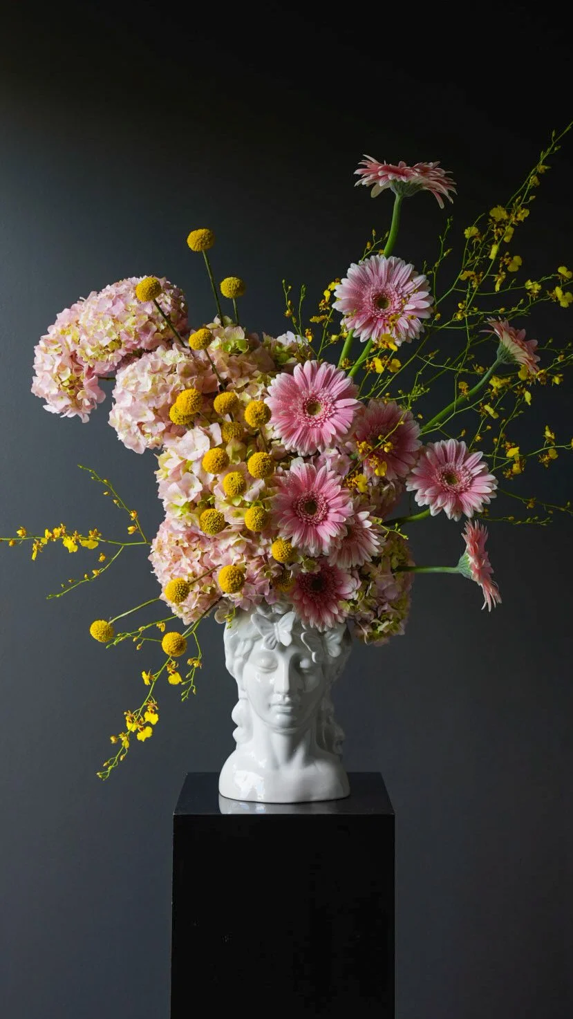A floral arrangement with pink and yellow flowers in a white sculpture vase shaped like a human face, placed on a black pedestal against a dark wall.