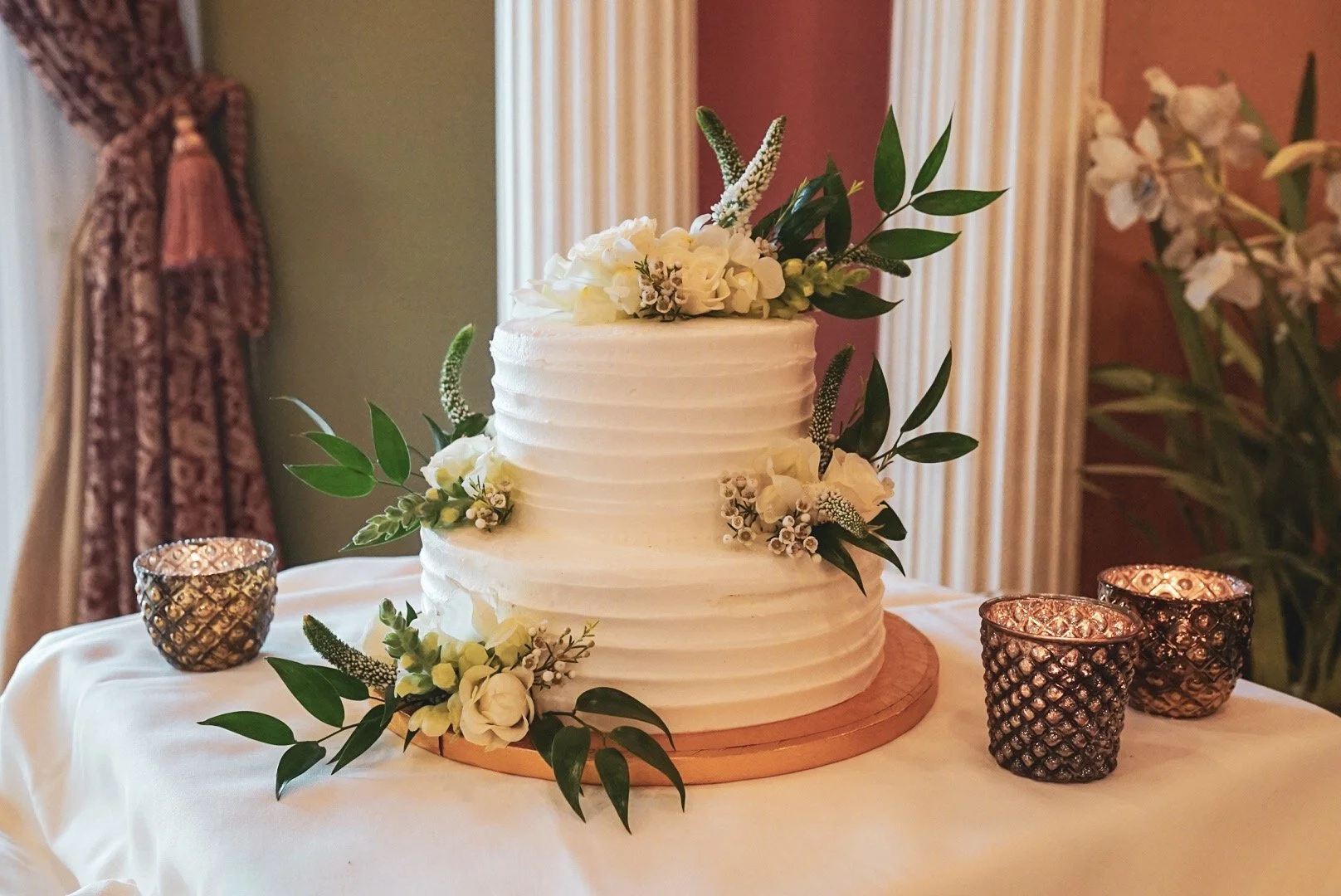 White wedding cake decorated with white flowers and green leaves, placed on a table with three lit candles in glass holders