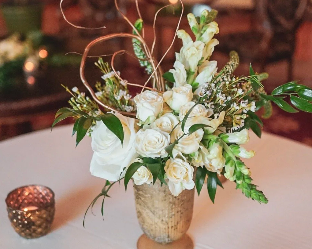 A floral arrangement of white roses, freesias, and greenery in a textured beige vase, placed on a round table with a small candle holder nearby.