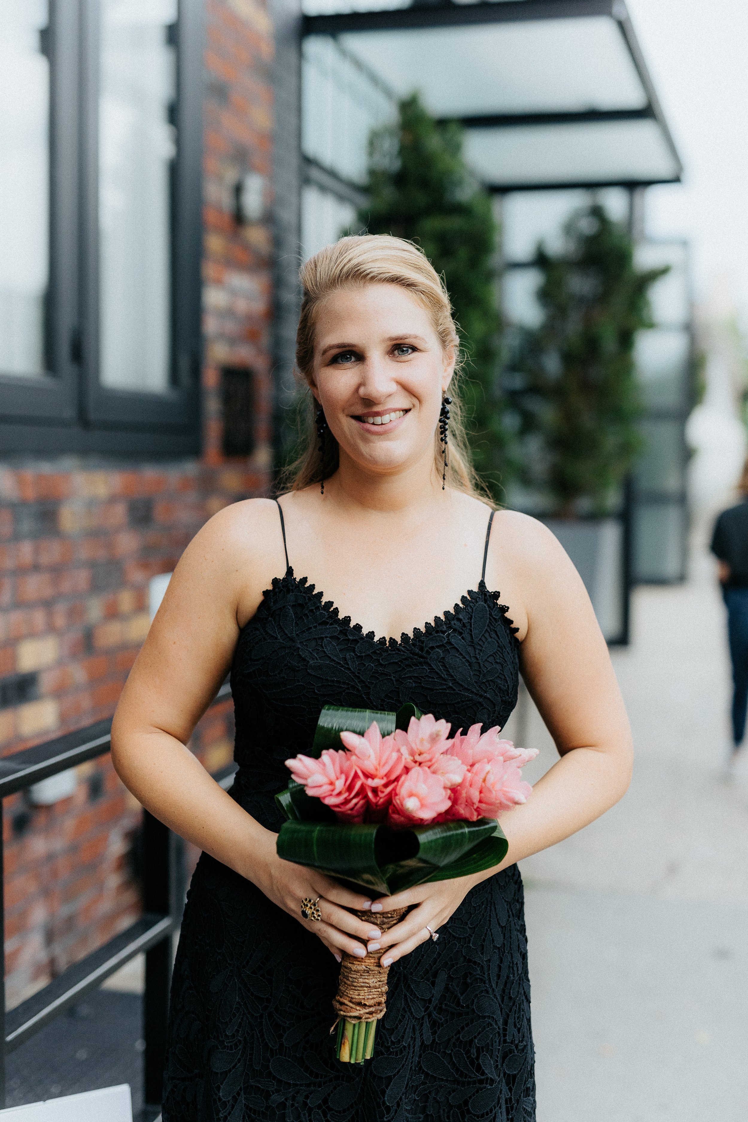 A smiling woman in a black lace dress holding a bouquet of pink flowers, standing outdoors on a city sidewalk.