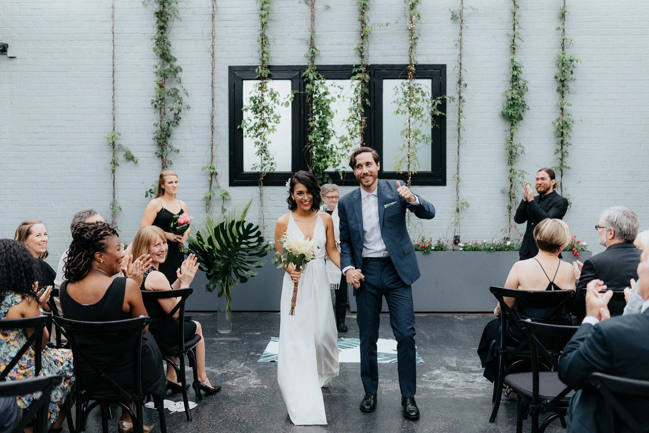 A newlywed couple walking down the aisle at their wedding, holding hands and smiling. The bride is holding a bouquet, wearing a white dress, and the groom is in a blue suit. Guests are clapping and celebrating around them inside a modern venue with g