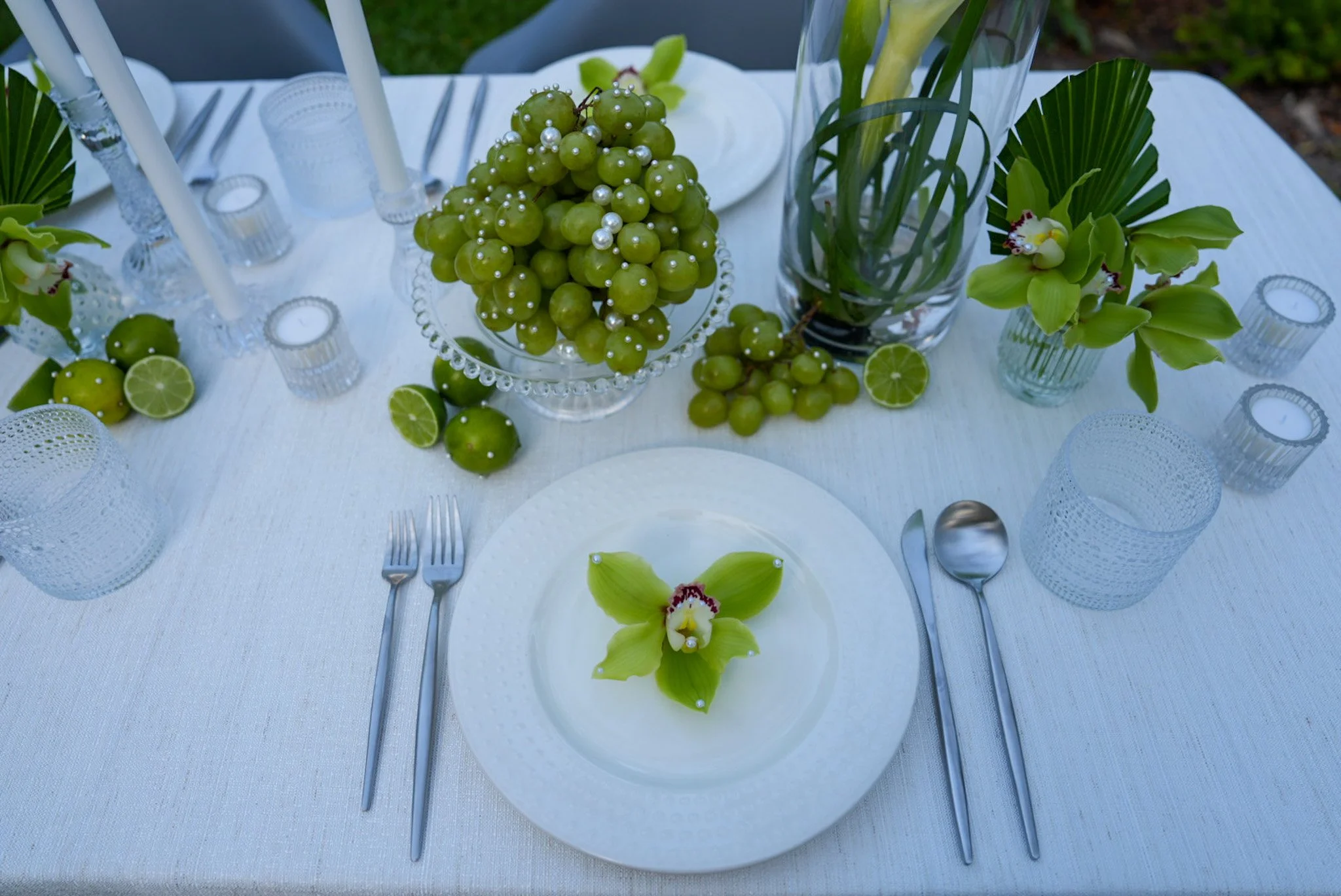 Table setting with white plates, silverware, glass tumblers, and decorative centerpieces including green grapes with pearl accents, lime halves, white orchids, and green leaves in glass vases on a white tablecloth.
