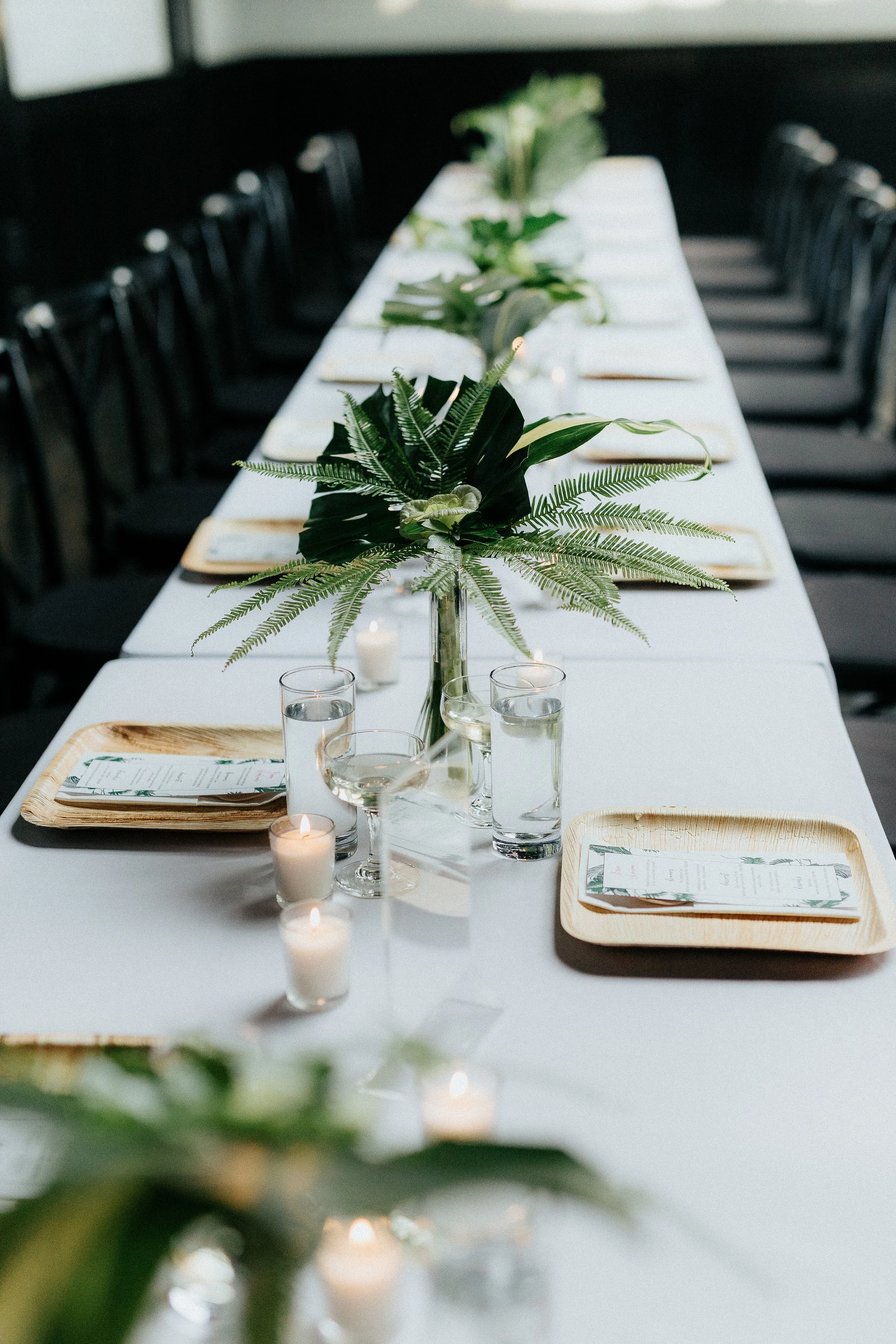 A long dining table decorated with greenery, candles, water glasses, and place settings with napkins and menus, set for a formal event.