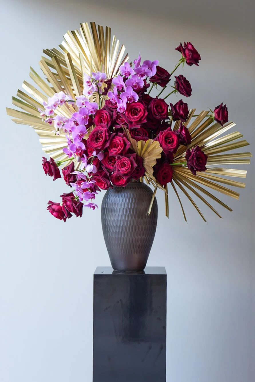 A floral arrangement featuring pink orchids and dark red roses in a textured gray vase, set on a black pedestal with gold decorative palm leaves in the background.