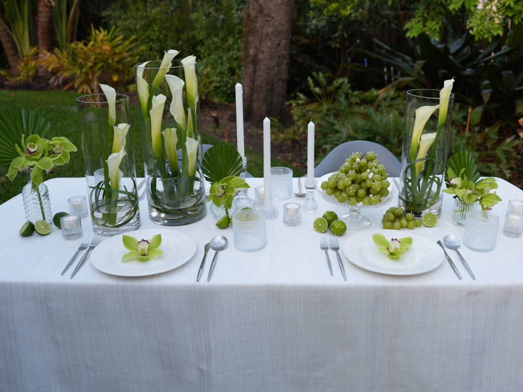 Elegant outdoor dining table decorated with white calla lilies, green orchids, grapes, candles, and white plates set for a meal.