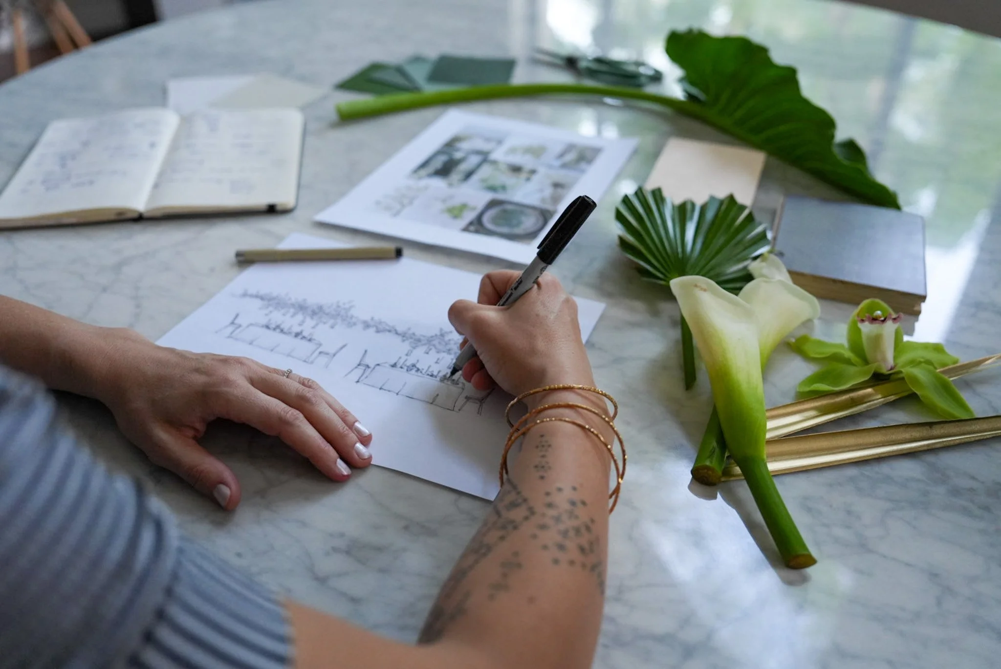 A person is sketching or drafting a landscape scene on a sheet of paper with a black marker, surrounded by tropical leaves and flowers on a marble table. The table also has notebooks, printed images, a pen, and a woman’s tattooed arm with gold bracelets.