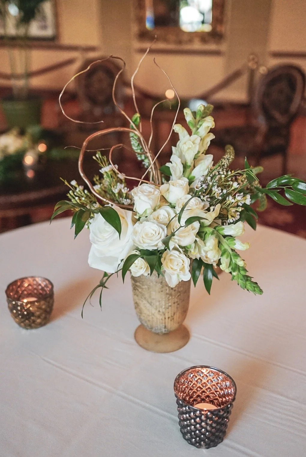 A floral arrangement with white roses and other white flowers, accented with green leaves, in a tall textured beige vase on a round table, with two small metallic candle holders nearby. In the background, a cozy room with wooden furniture and a window.