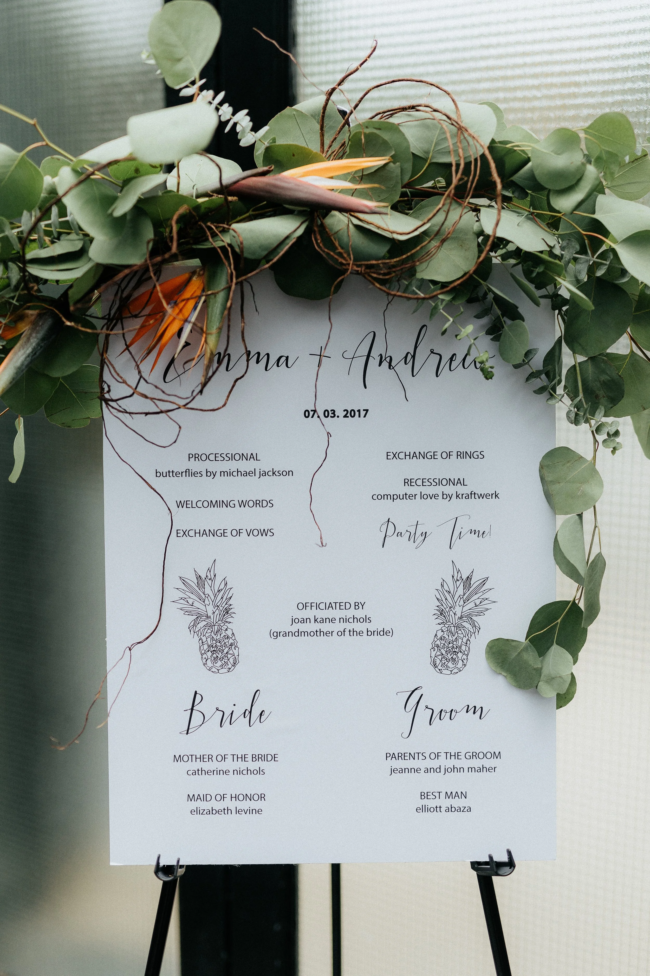 Wedding program sign on easel decorated with green leaves and eucalyptus, listing the ceremony details and wedding party members.