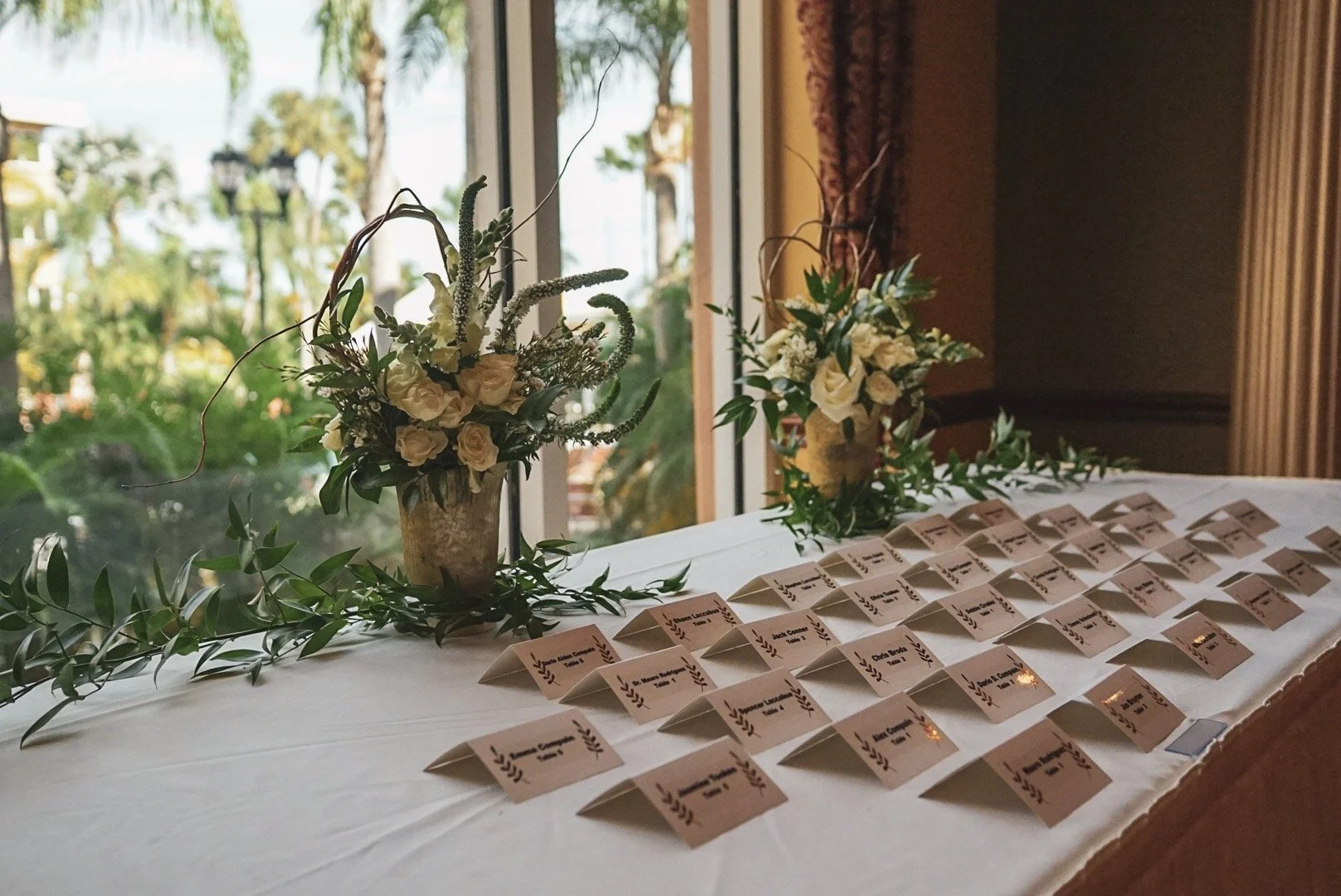Table with a white tablecloth, decorated with two floral arrangements in vases, and arranged name cards, near a window with a view of trees outside.