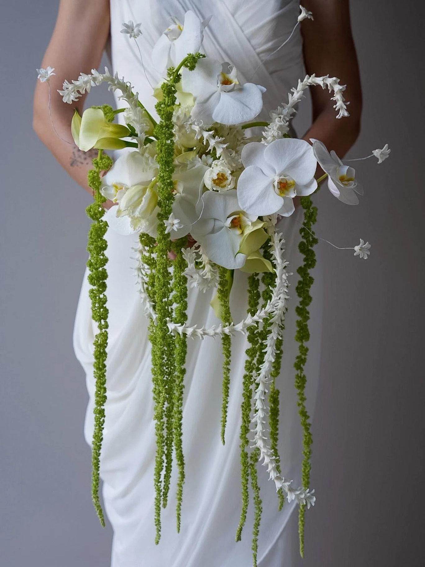 A person in a wedding dress holds a cascading bridal bouquet of white orchids and green hanging amaranthus.