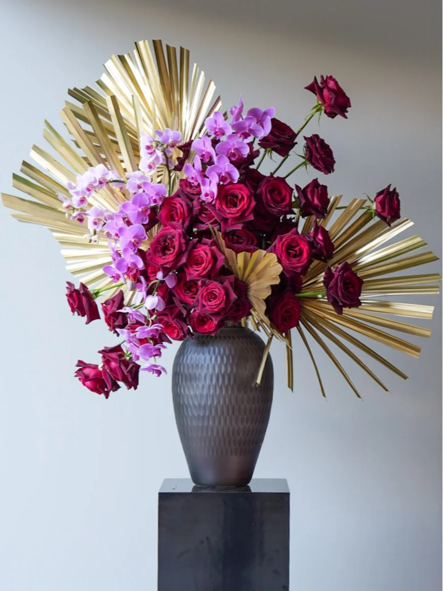 A flower arrangement with pink orchids and red roses in a textured gray vase, accented with large gold decorative leaves, on a black pedestal against a light background.