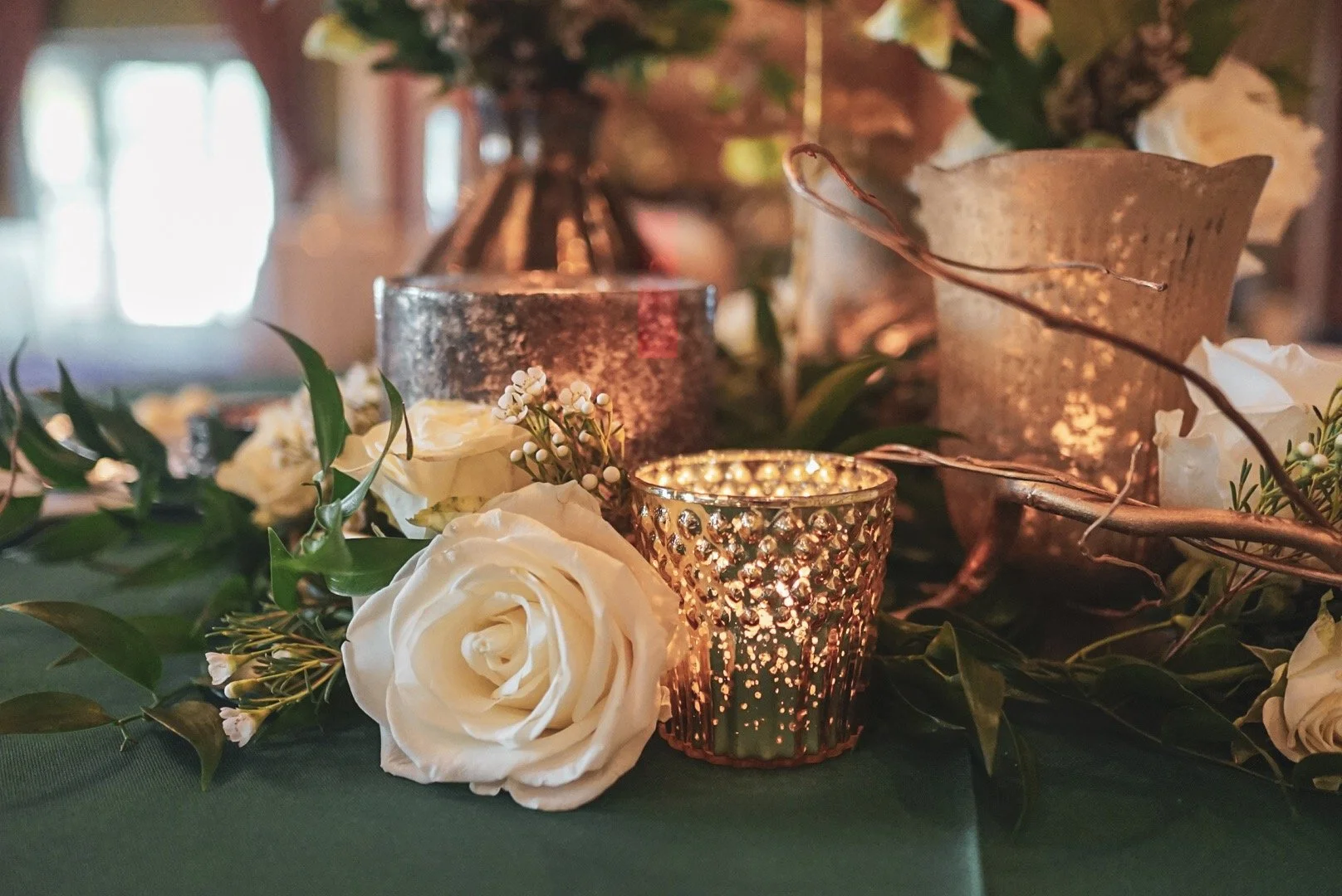 A close-up of a table centerpiece with white roses, green leaves, small white flowers, and lit candles in decorative holders at an event.