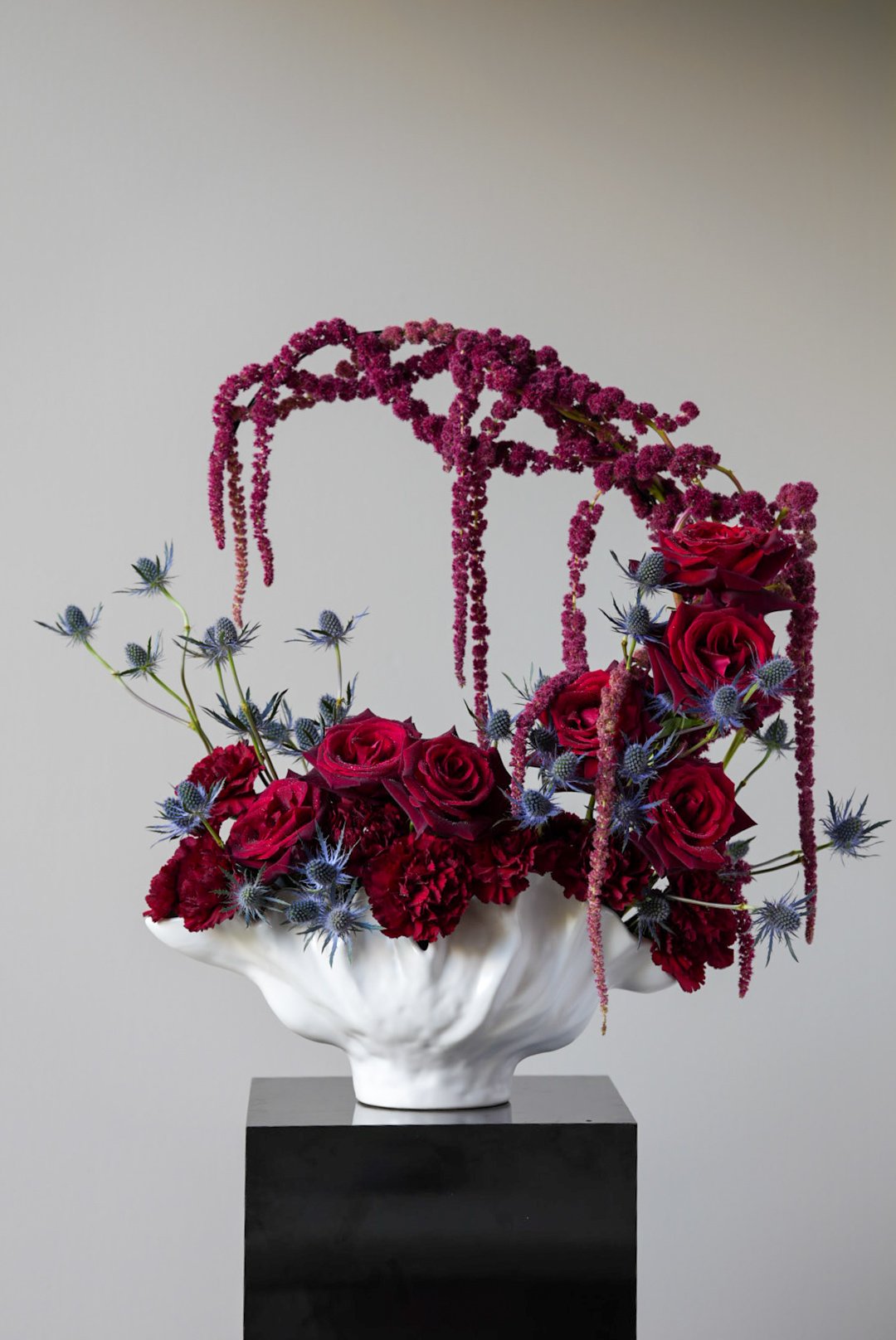 A floral arrangement featuring deep red roses, red carnations, purple thistle, and hanging purple amaranth, displayed in a white ceramic bowl on a black pedestal against a plain background.