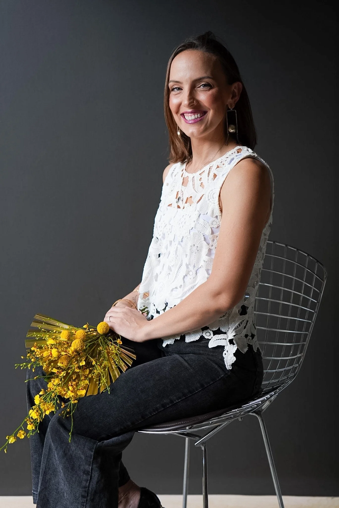 A woman with shoulder-length brown hair and earrings, smiling, wearing a white lace sleeveless top and black jeans, sitting on a modern wire chair against a dark gray background, holding a bouquet of yellow flowers.