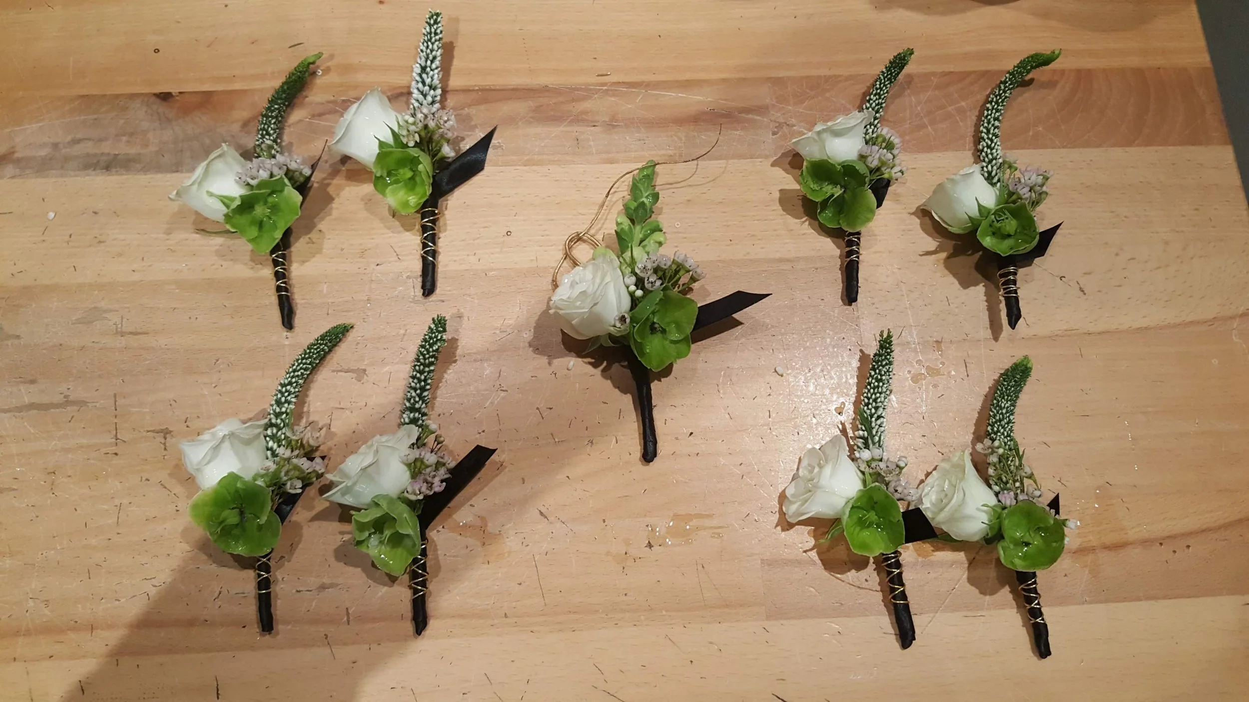 Eight floral boutonnieres arranged on a wooden table, each featuring a white rose, green leaves, and small white and purple flowers, with black stems wrapped in gold wire.