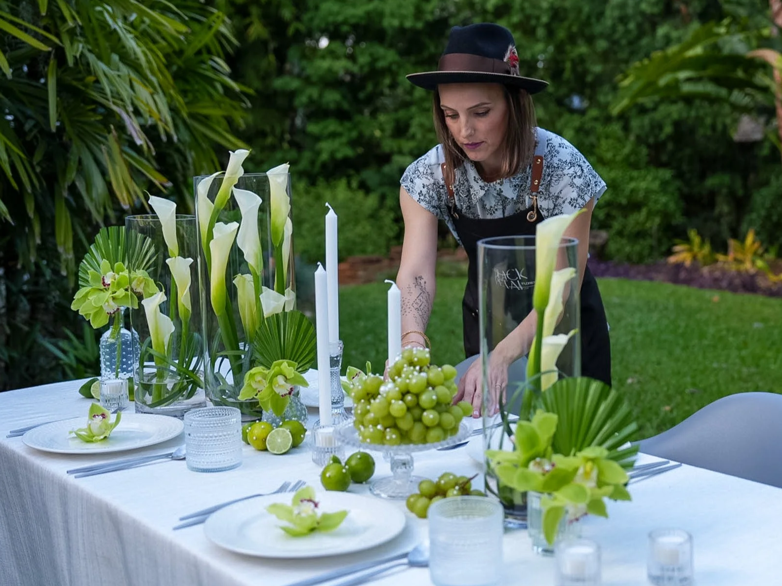 A woman arranging a table centerpiece outdoors with white calla lilies, green grapes, and white candles in tall glass vases, surrounded by greenery.