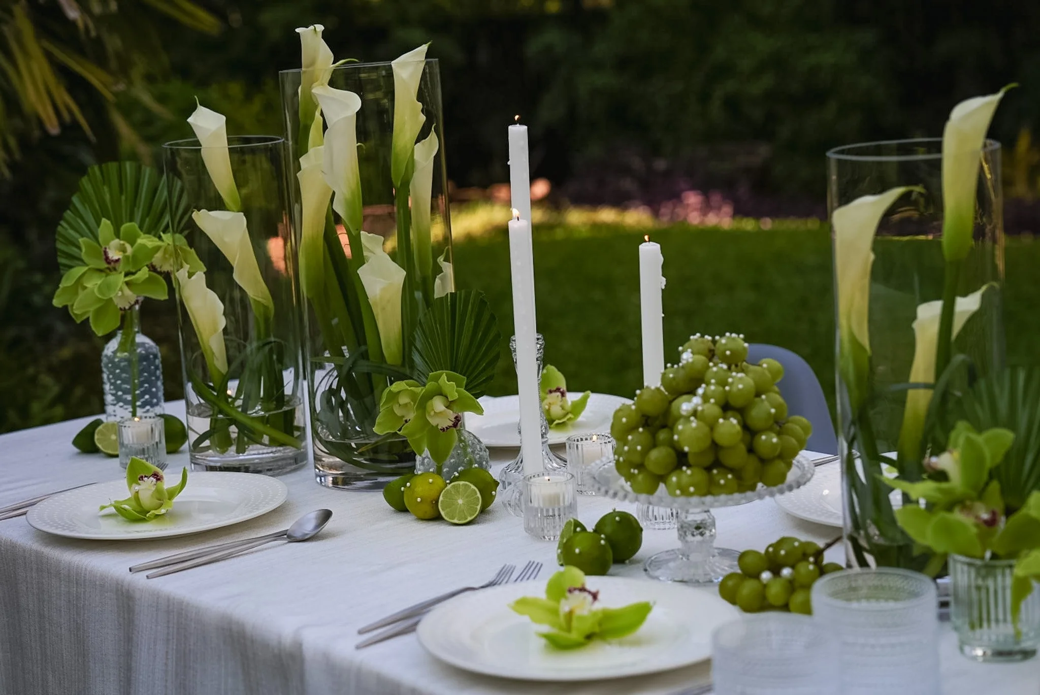Elegant outdoor table setting with white tablecloth, floral arrangements in tall glass vases, white candles, and a centerpiece featuring green grapes, lime slices, and flowers, with a grassy background.