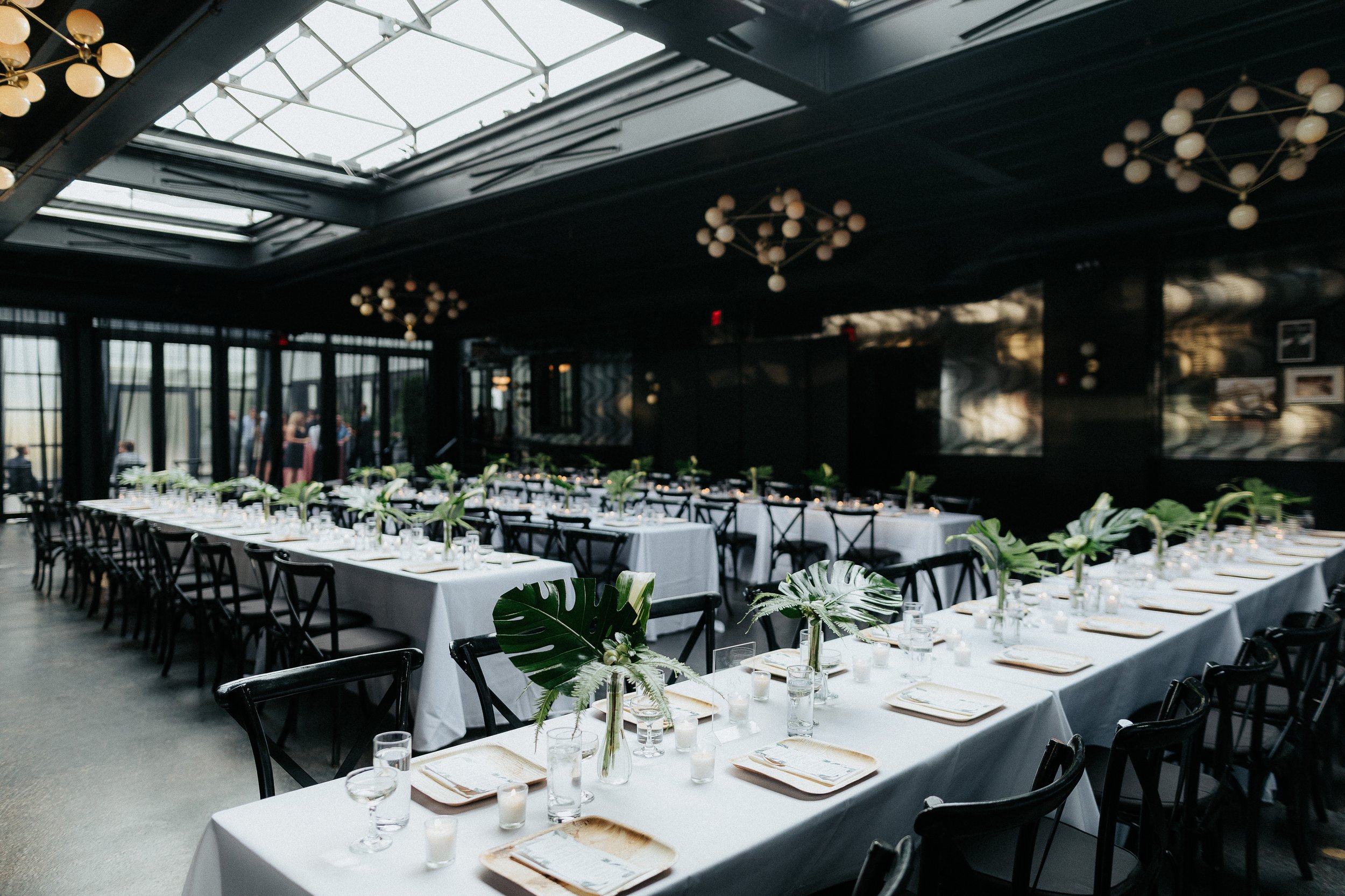 Elegant indoor event space with long tables set for a formal gathering, decorated with large tropical leaves in glass vases, white tablecloths, glassware, and candles, with modern black chairs and chandelier lighting.
