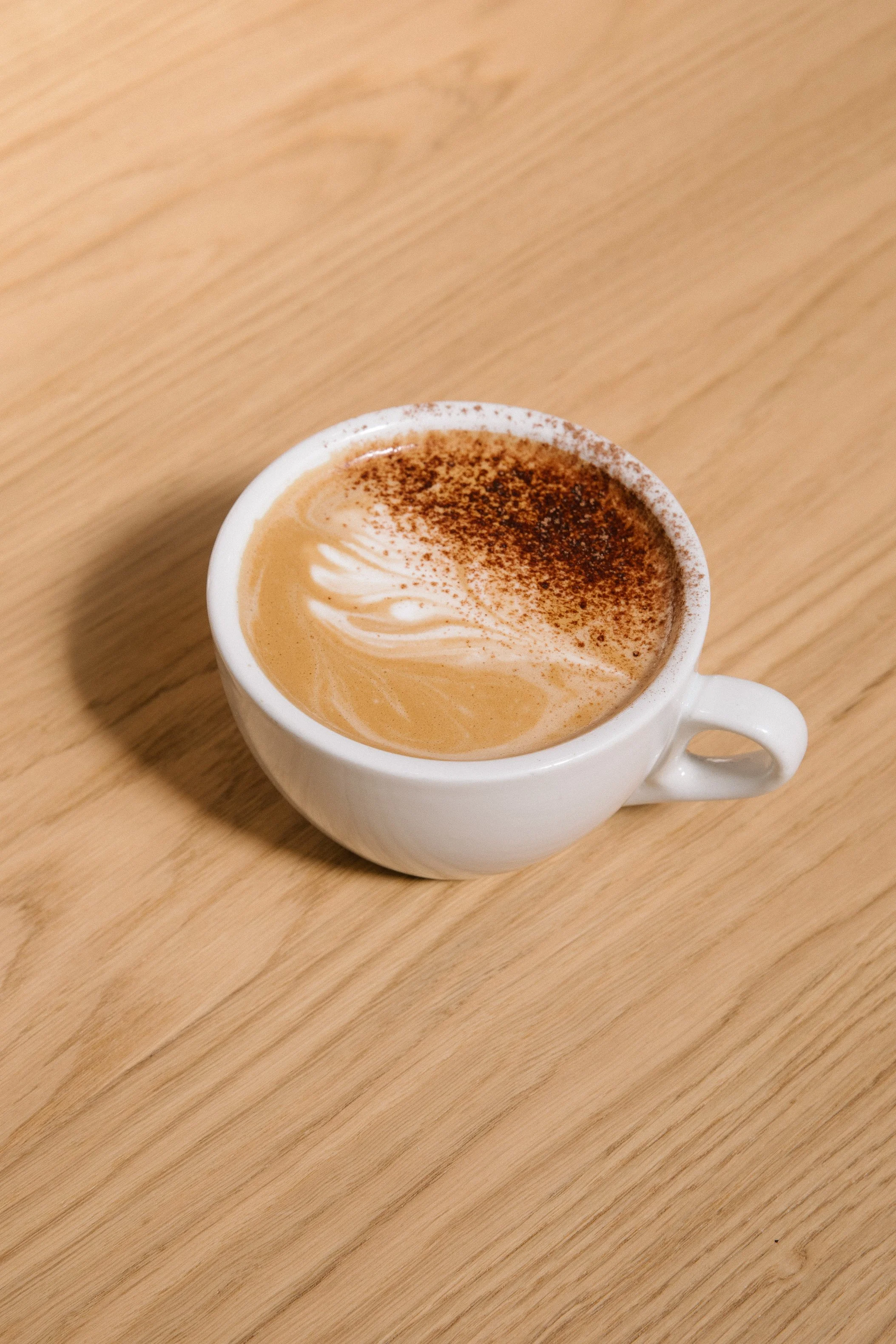 A white ceramic cup of cappuccino with cinnamon sprinkled on top, placed on a wooden table.
