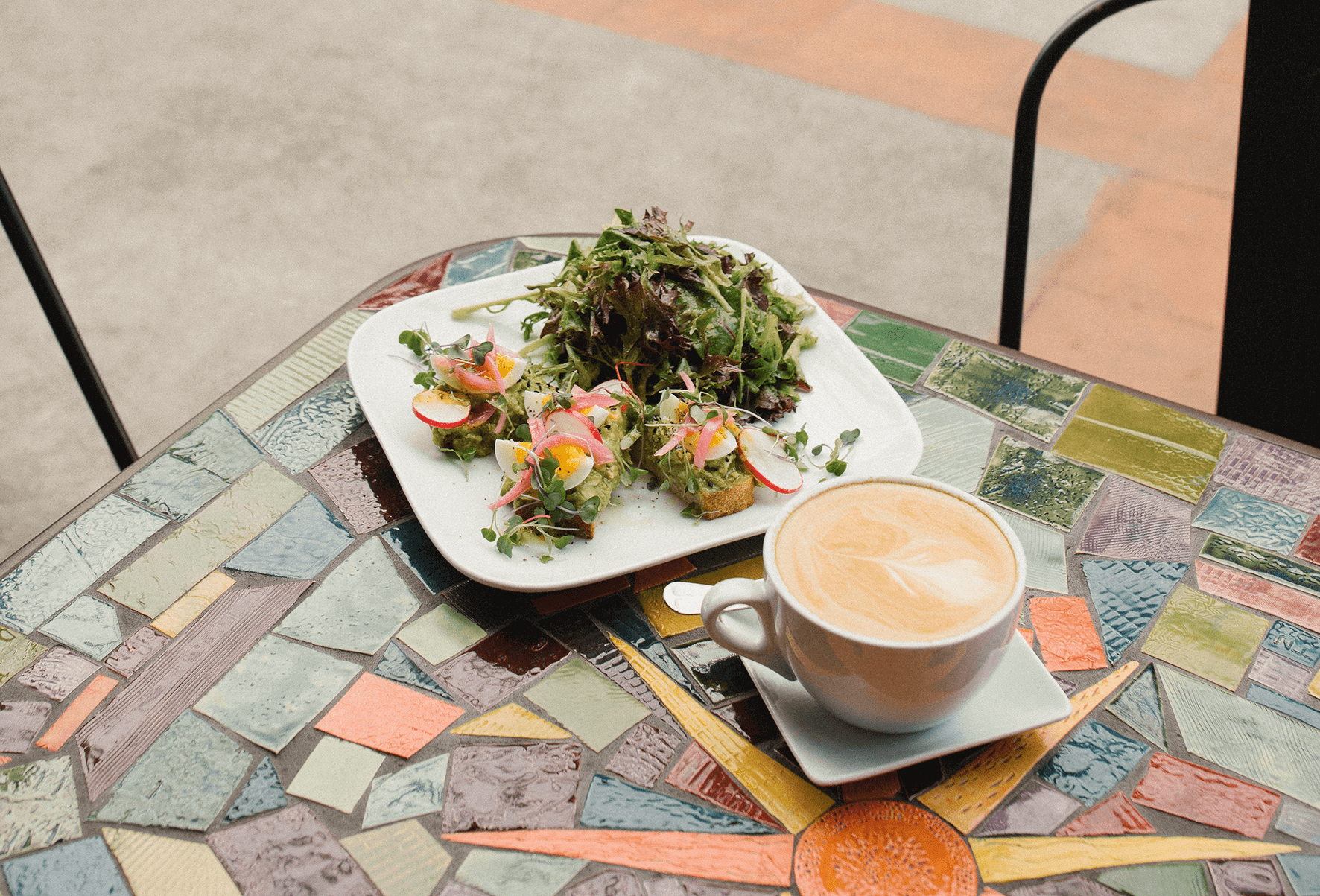 A table with a colorful mosaic tile surface holds a white plate of avocado toast and salad with mixed greens, radish slices, and microgreens, and a cup of coffee with a saucer.