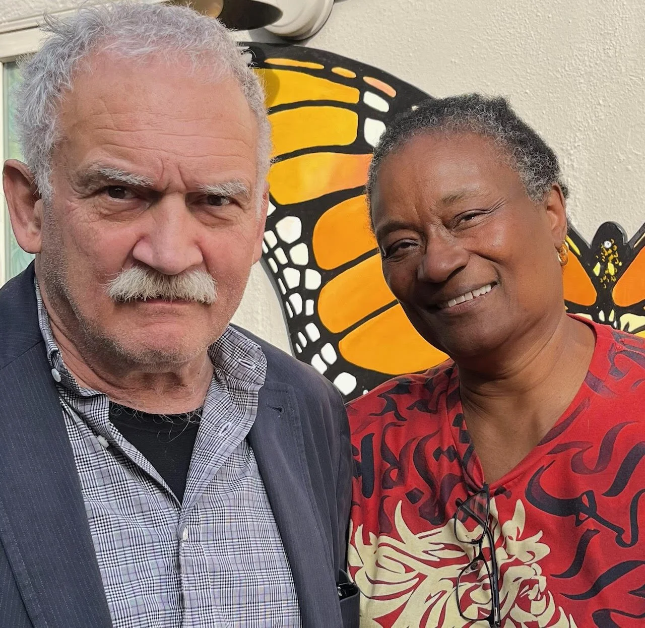 Close-up photo of an elderly white man with gray hair and a white mustache, wearing a plaid shirt and blazer, standing next to an elderly woman with dark skin and short gray hair, wearing a red shirt with a dragon print, both smiling in front of a butterfly artwork with orange and black wings.