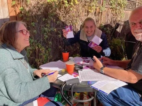 Three people sitting around a table outdoors, smiling and holding cards, with papers and drinks on the table.