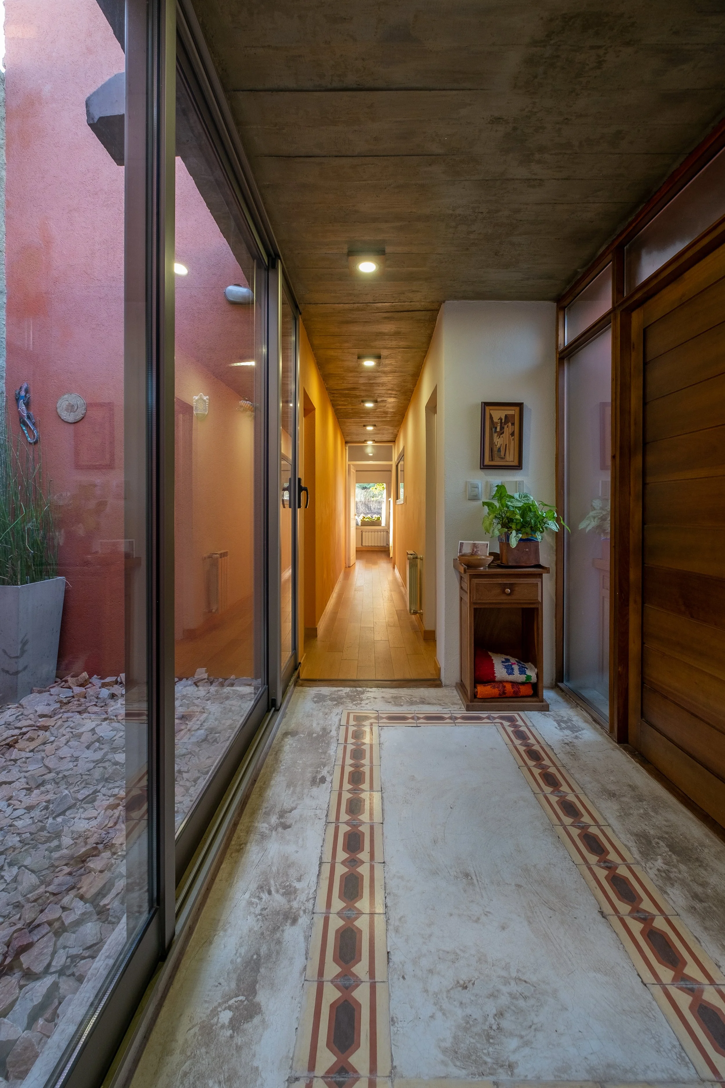 Vista de un pasillo con muros en madera y tonos amarillos, cielorraso de hormigón con iluminación empotrada, una pequeña mesa lateral de madera con una planta y objetos decorativos, grandes puertas corredizas de vidrio, casa en Alta Gracia, Córdoba.