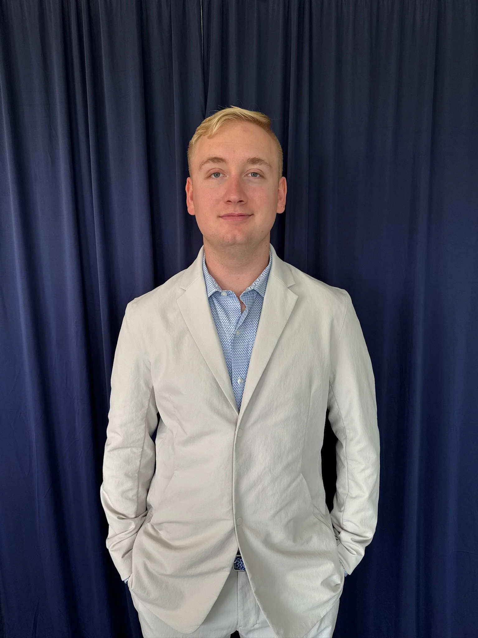 A young man in a light-colored suit jacket and blue patterned shirt standing in front of navy blue curtains.