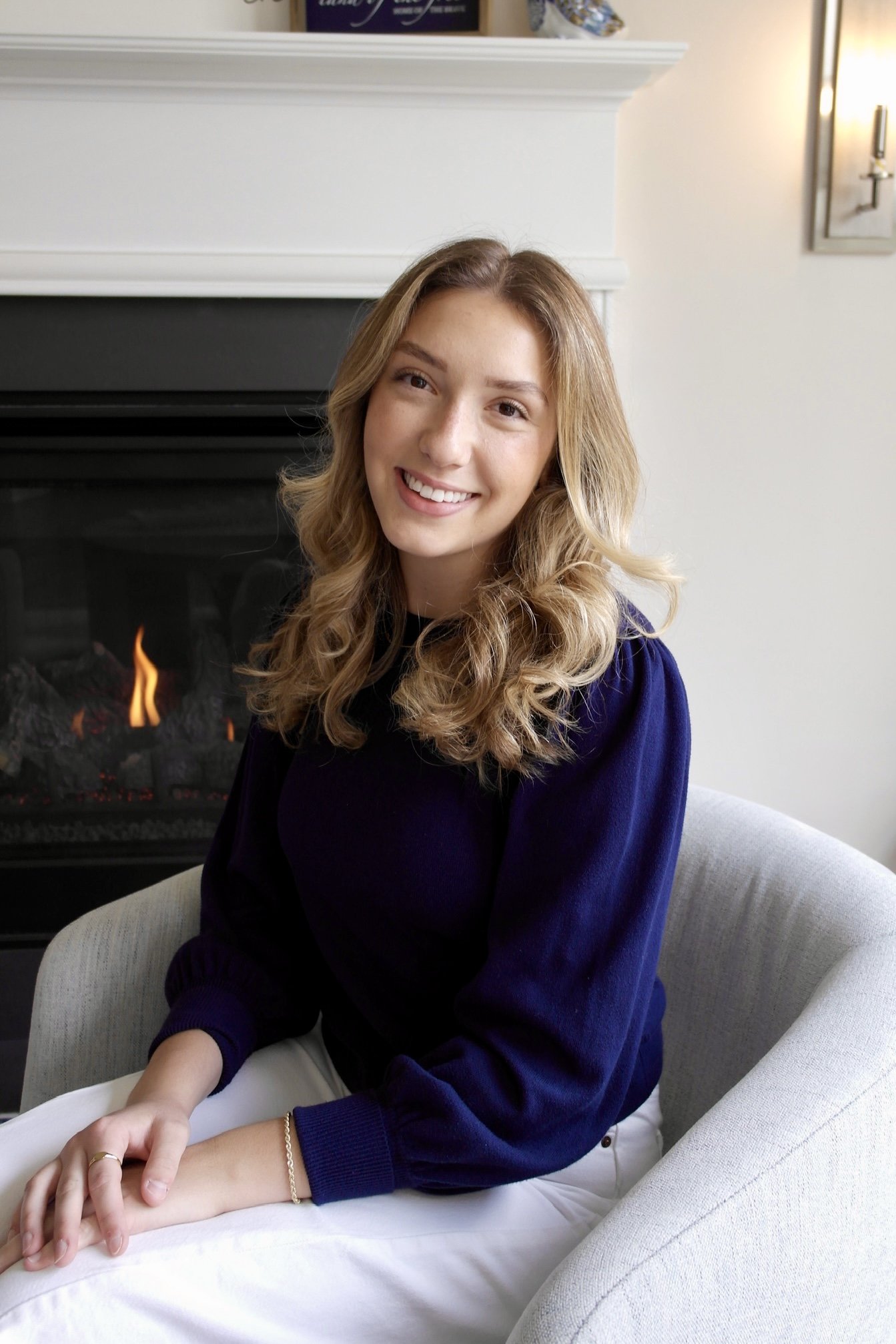 A young woman with blonde curly hair and a big smile sitting on a beige chair, wearing a navy blue sweater and white pants, in a room with a fireplace in the background.