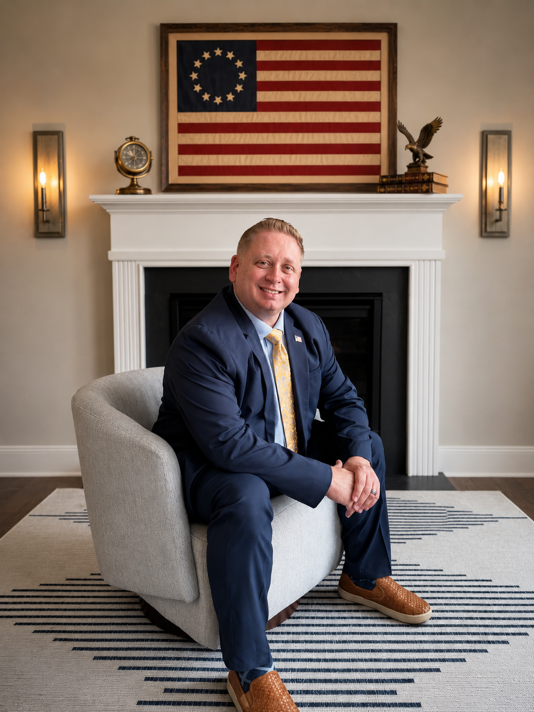 A man in a navy suit with a yellow tie sitting on a light gray armchair in front of a white fireplace with an American flag-themed artwork hanging above it. The room has wall-mounted lights and decorative items including a clock and an eagle sculpture.