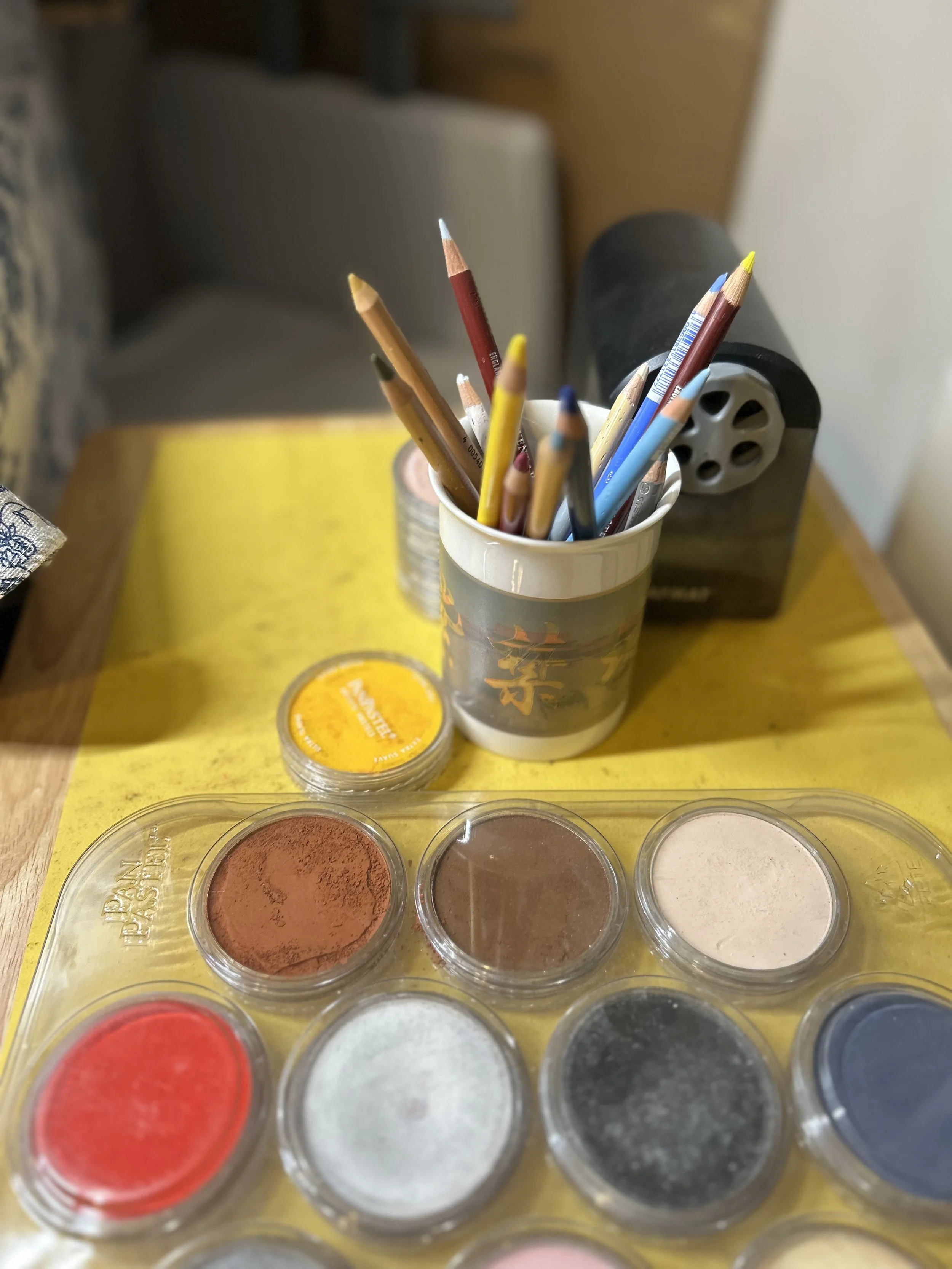 A desk with a yellow placemat, a small container of pigment powders, a cup with colored pencils, and a tape dispenser.