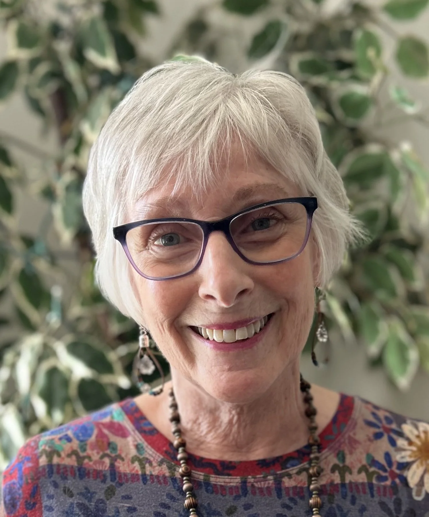 A smiling elderly woman wearing glasses, earrings, and a colorful patterned top with a necklace, standing in front of a leafy green plant.