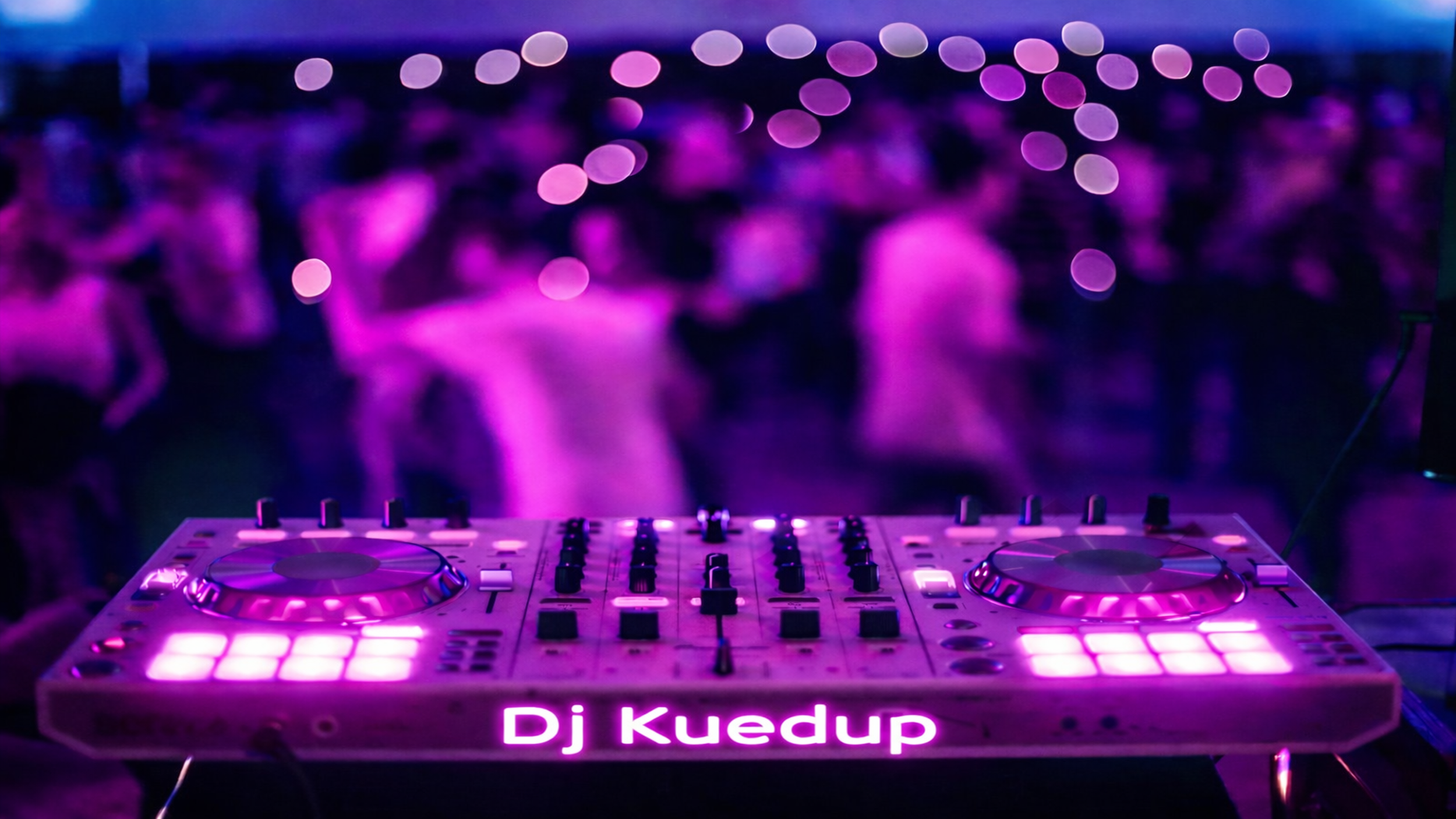 A DJ set up with glowing purple lights and the name 'Dj Kuedup' illuminated at the front, with a blurred crowd of people dancing in the background at a nightclub or party event.