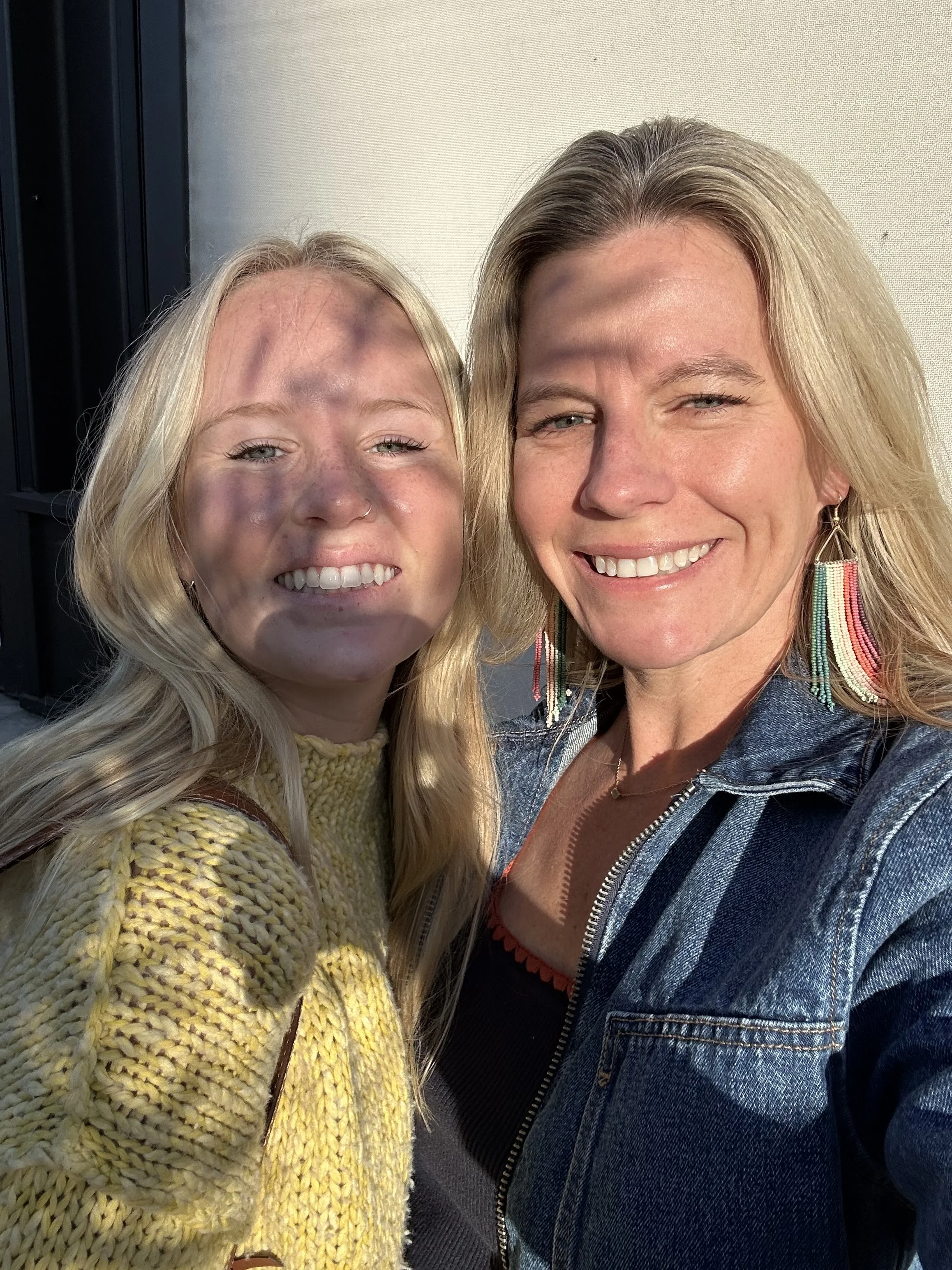 Two women taking a selfie together outside, smiling, with sunlight on their faces. One has blonde hair, the other has slightly darker blonde hair, and both are wearing earrings.