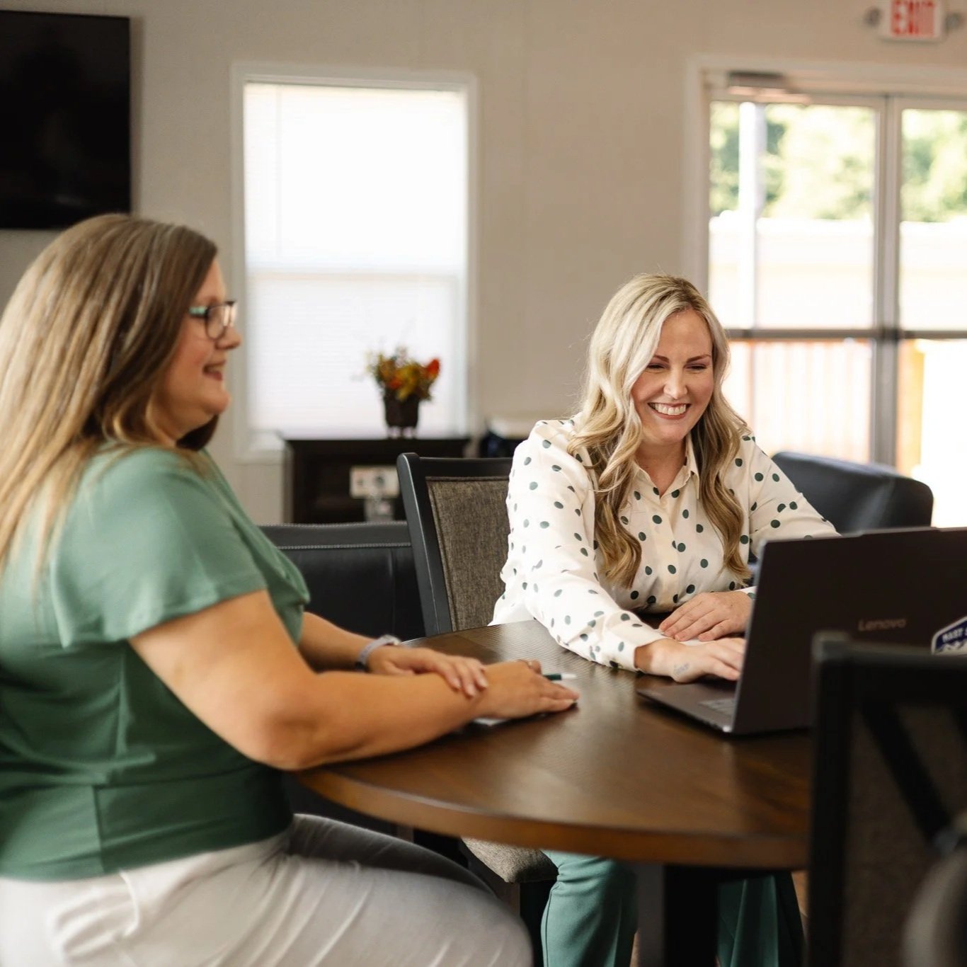 Two women sitting at a conference table, smiling and looking at a laptop. One woman has long blonde hair and is wearing a white blouse with black polka dots. The other woman has long brown hair and is wearing glasses and a green top.