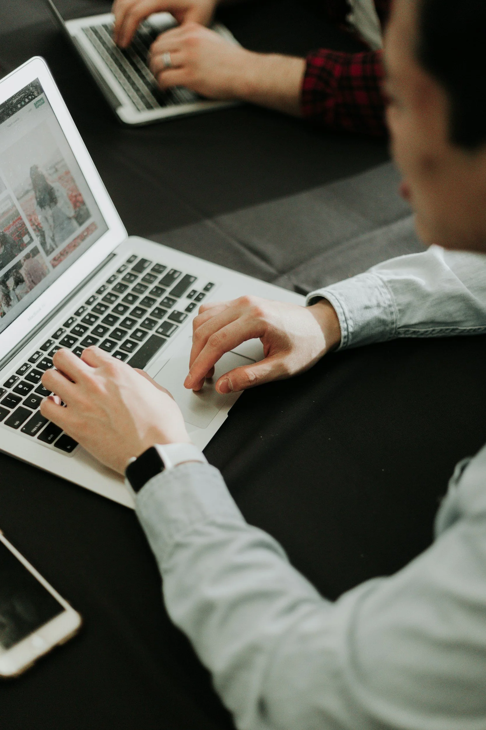 Person using a laptop with a smartphone on the table, another person working on a laptop in the background.