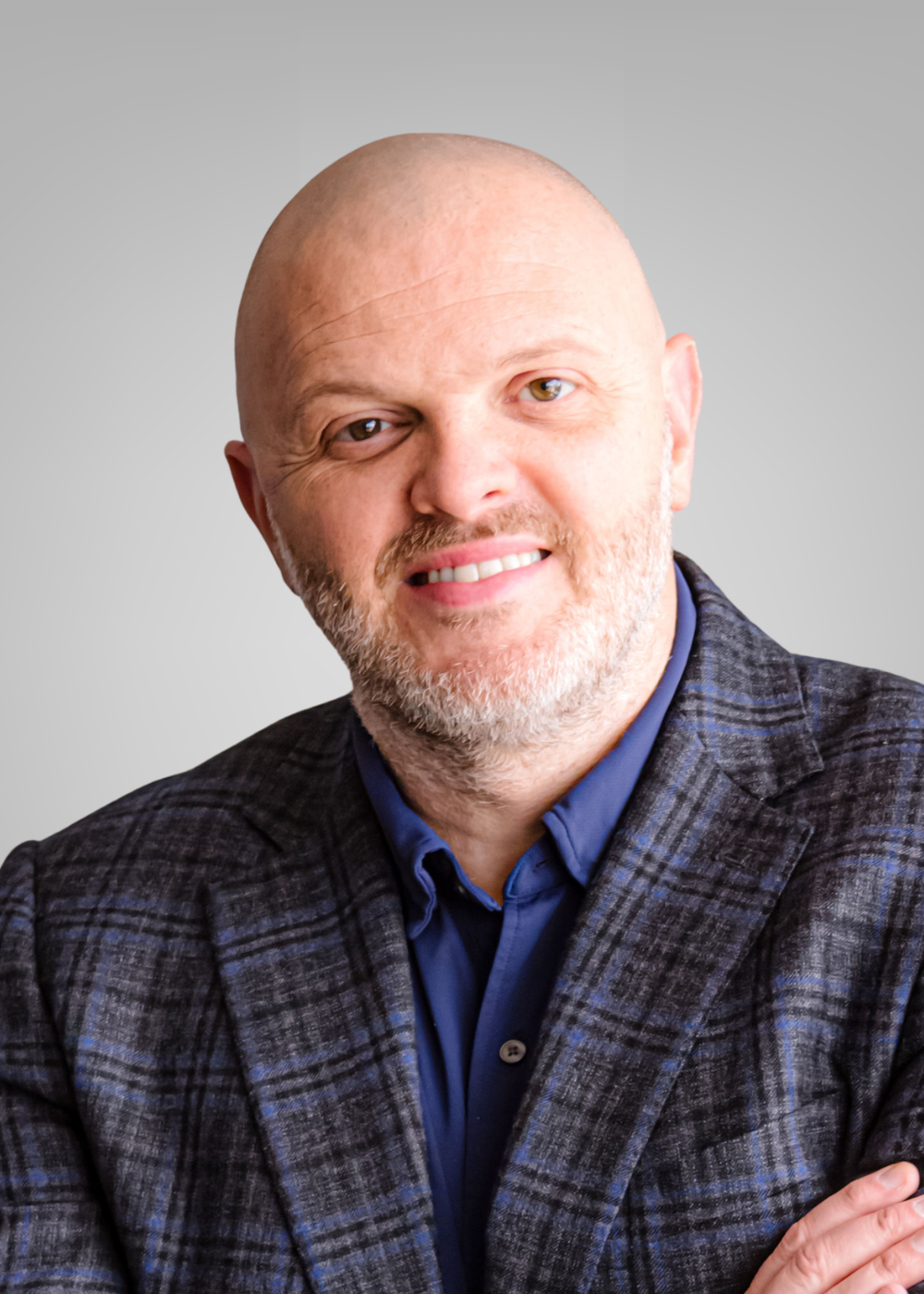 A man with a bald head, beard, and mustache wearing a dark plaid blazer and blue shirt, smiling with arms crossed against a plain gray background.