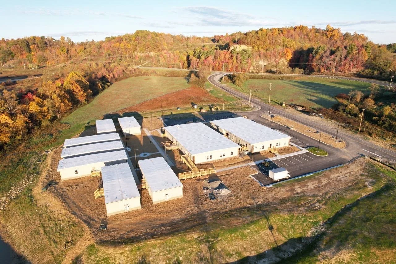 Aerial view of a newly built modular housing complex with several white buildings, parking lot, and surrounding green fields and forested hills.